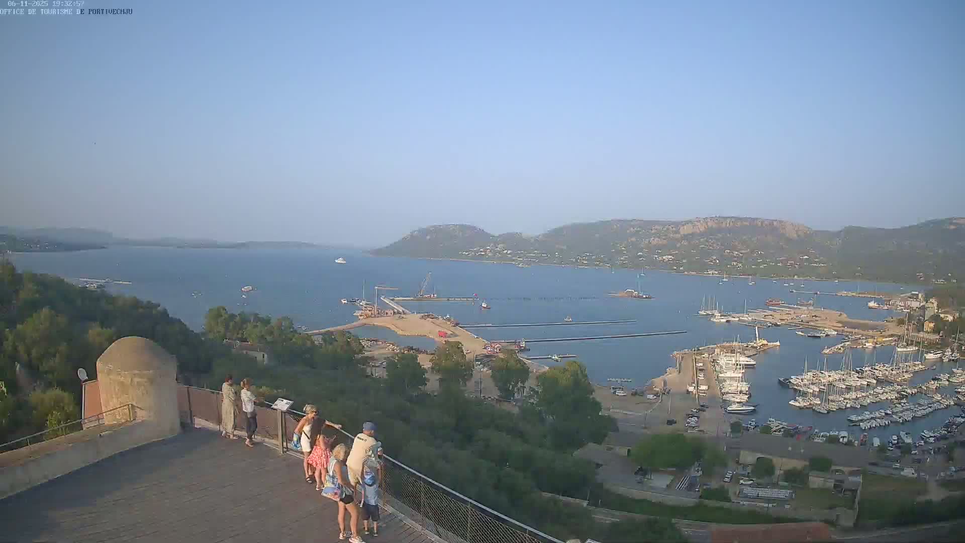 A clear, sunny day overlooking a harbor filled with numerous boats and yachts, with several people standing on a viewing platform in the foreground.