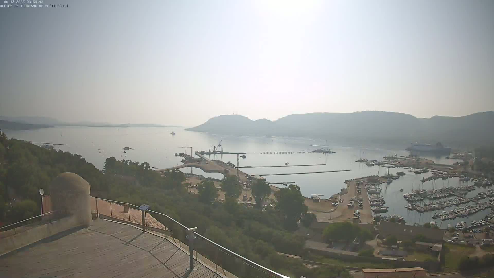 A hazy, sunny day overlooks a harbor filled with numerous boats and yachts, situated near a shoreline with green vegetation and hills in the background.