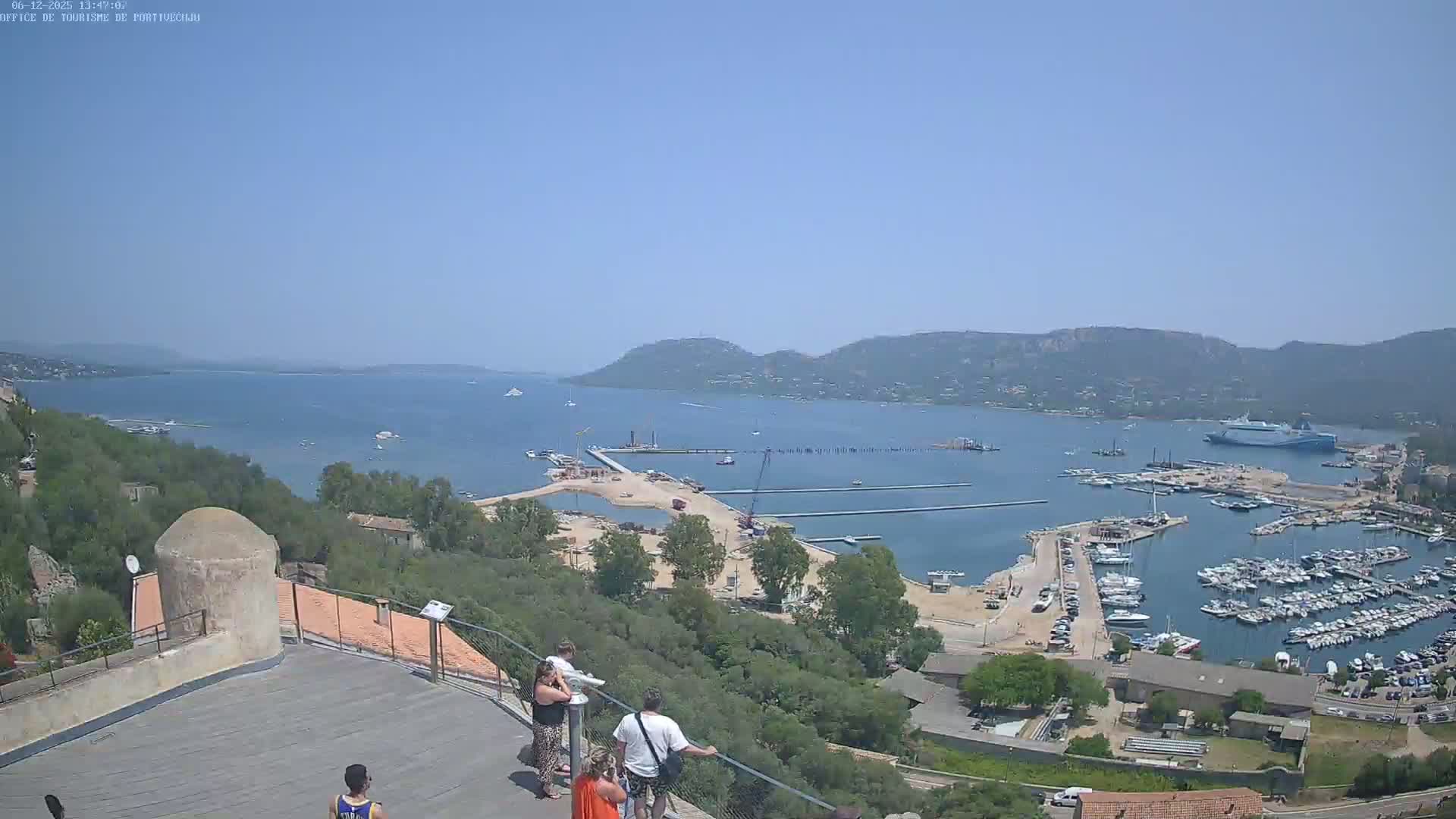 An elevated view on a clear, sunny day shows a harbor filled with boats, a partially developed waterfront area, and a calm bay surrounded by hills and vegetation, with several people looking out from a high vantage point.
