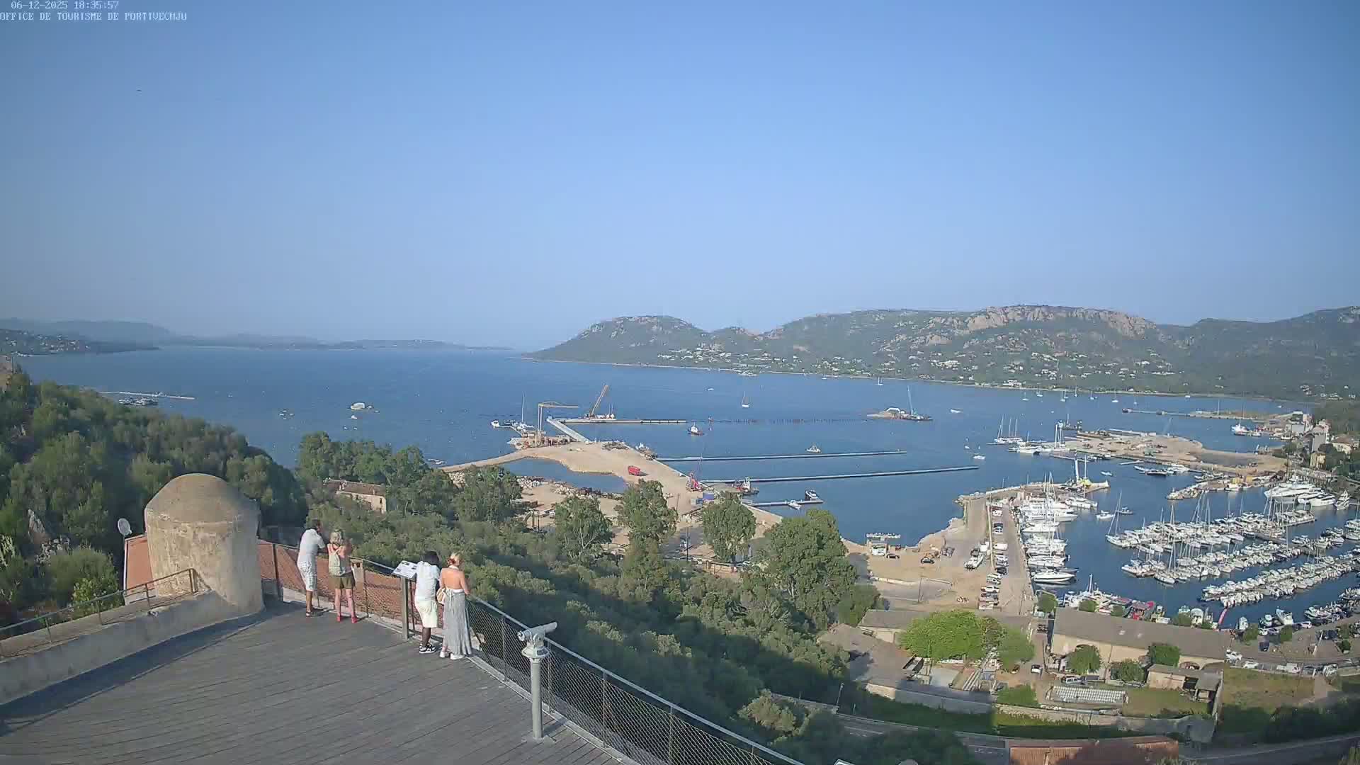 A high-angle, long shot shows a sunny day overlooking a harbor filled with boats, construction, and people standing on a viewing platform, with hills and a bay in the distance.