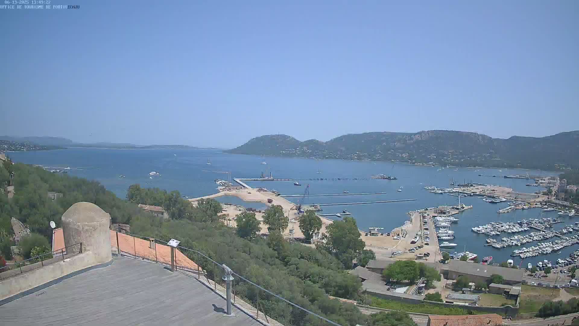 A high-angle, long shot on a clear, sunny day shows a harbor filled with boats, construction in progress on a nearby pier, and a picturesque coastal town nestled amongst green hills overlooking a calm bay.