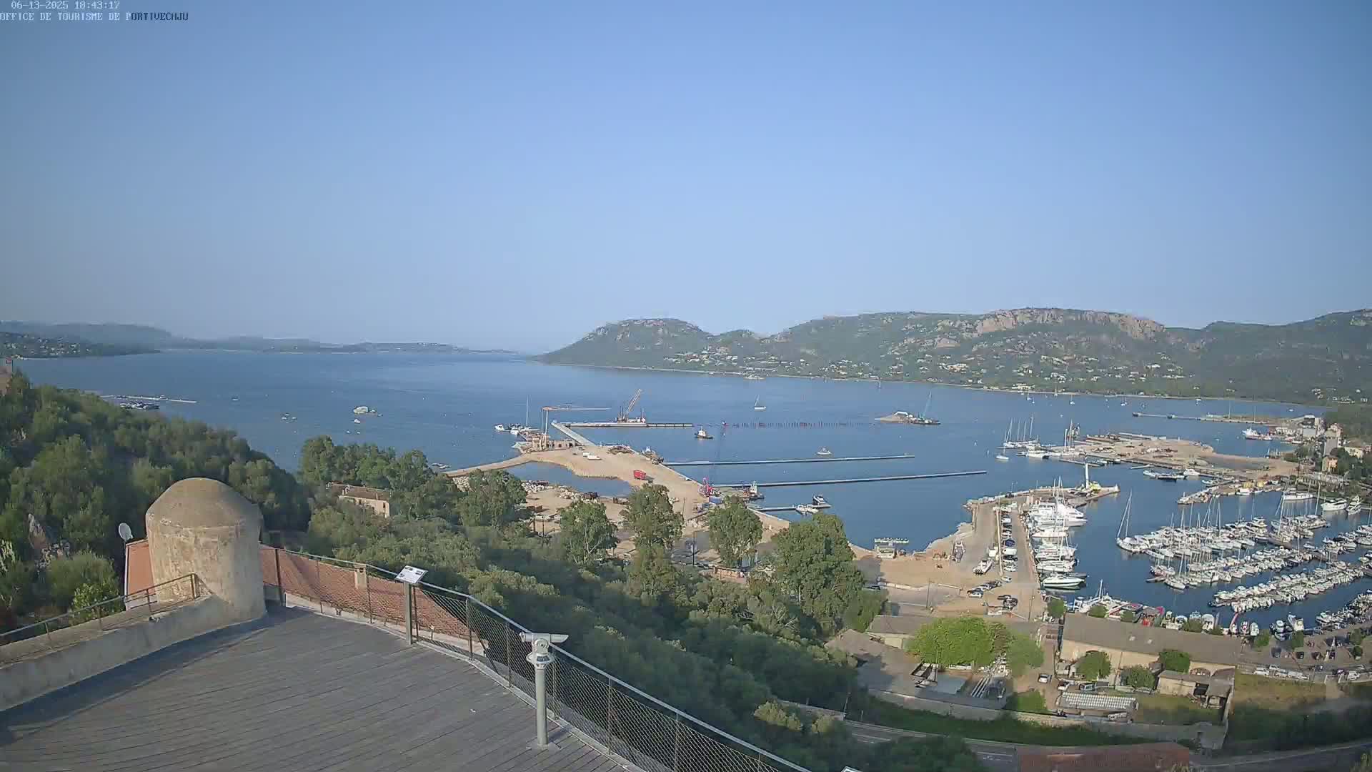 A high-angle view on a clear day shows a marina filled with boats, construction underway on a pier, and a calm bay extending to distant hills.