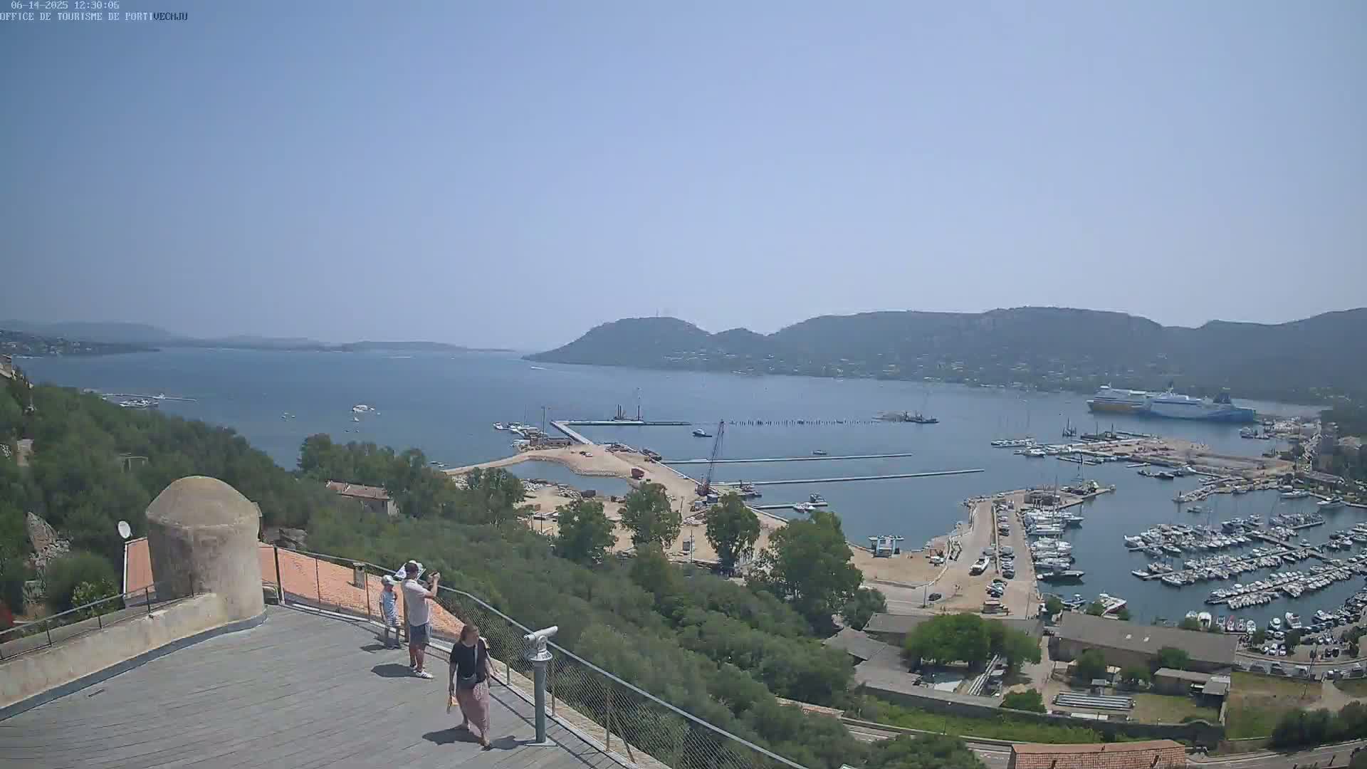 An elevated view shows a harbor filled with boats on a sunny day, with a large ferry in the background and several people standing on a rooftop overlooking the scene.