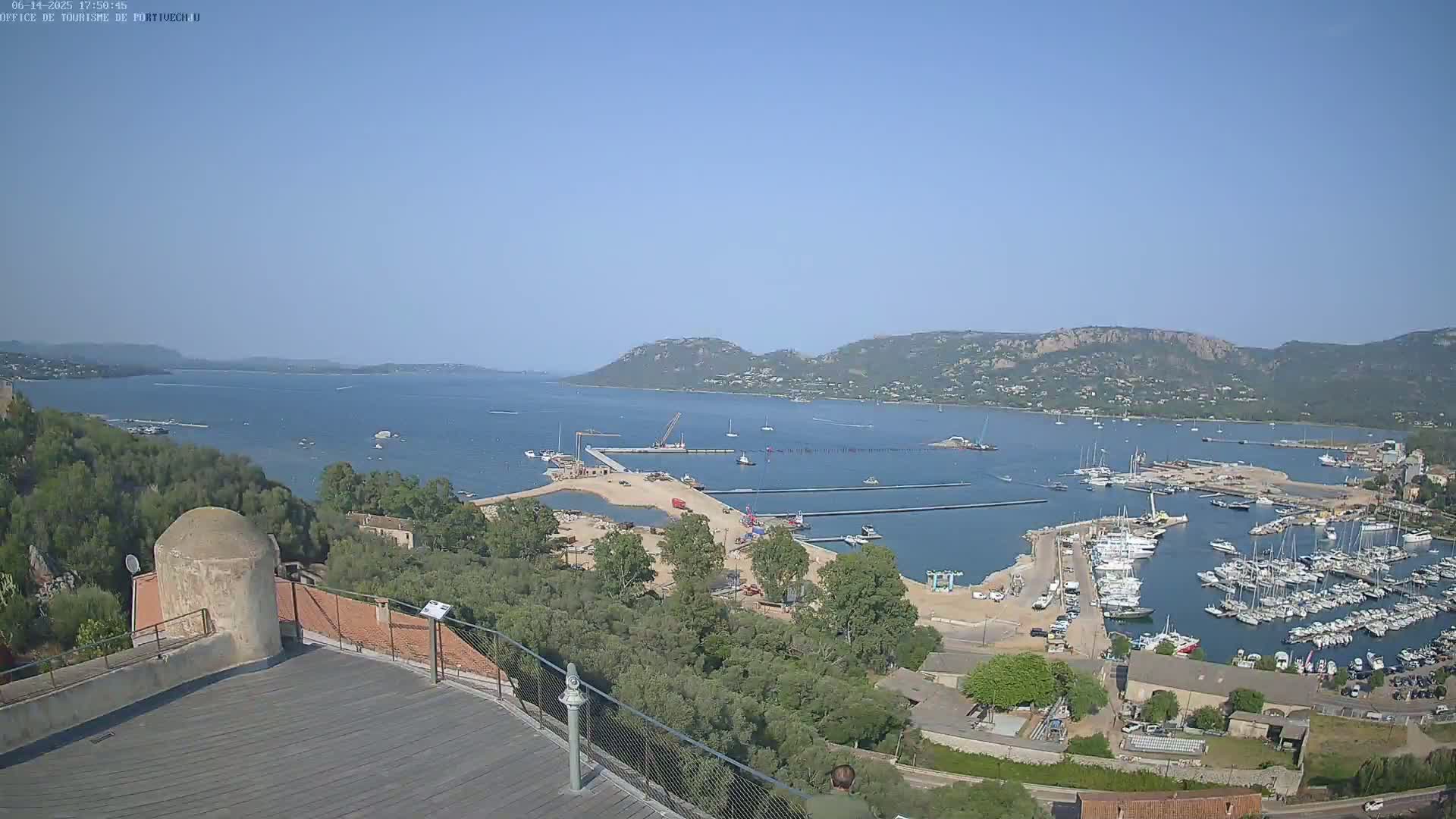 A high-angle, long shot on a clear, sunny day shows a harbor filled with boats, construction activity near a sandy area, and green hills in the background across a calm body of water.