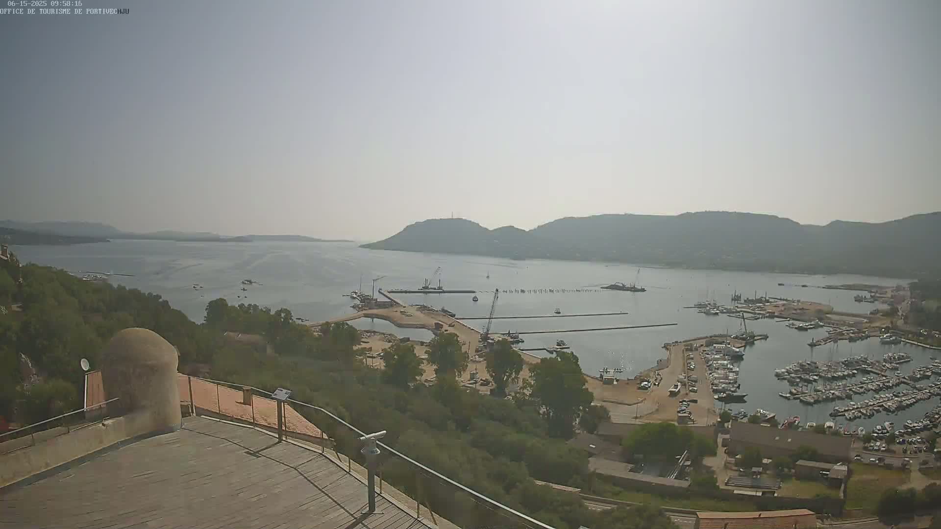 A hazy, sunny day overlooks a harbor with numerous boats docked, construction activity, and hills in the background, viewed from a high vantage point.