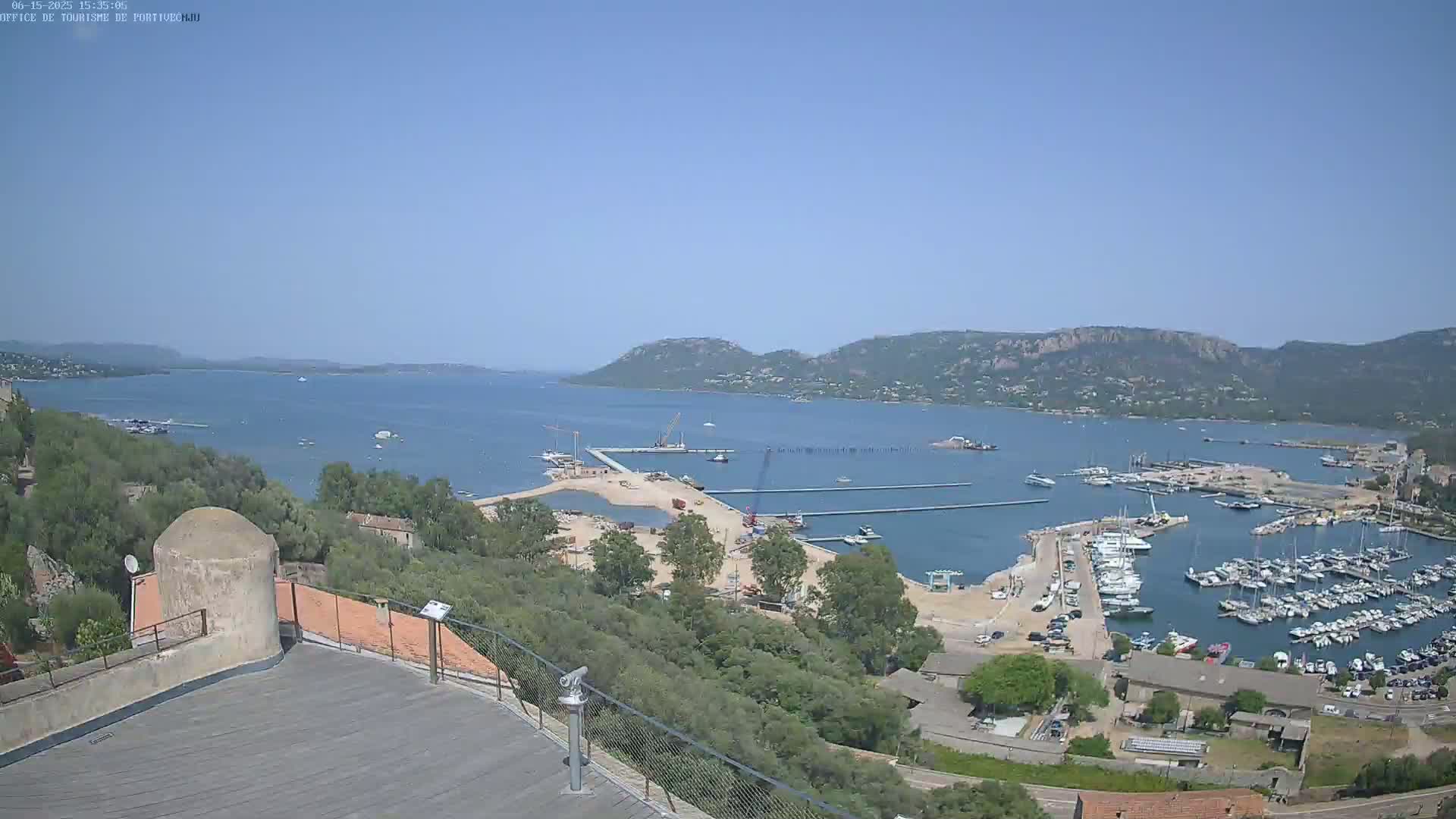 A high-angle, sunny day view shows a harbor filled with boats, construction on a nearby pier, and a bay surrounded by hills and trees.