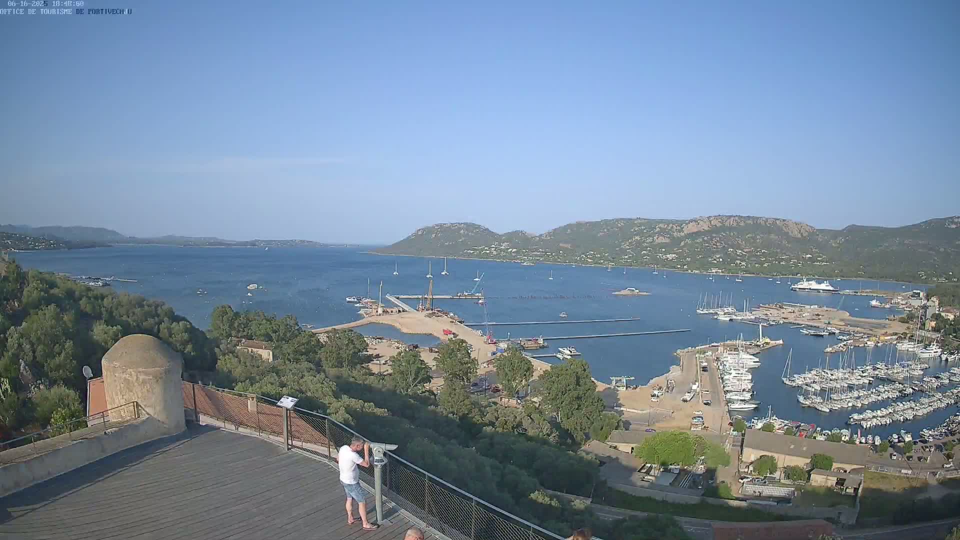 An elevated view of a harbor filled with boats, under clear blue skies, shows construction activity near a sandy beach with a person using a telescope on a nearby rooftop.