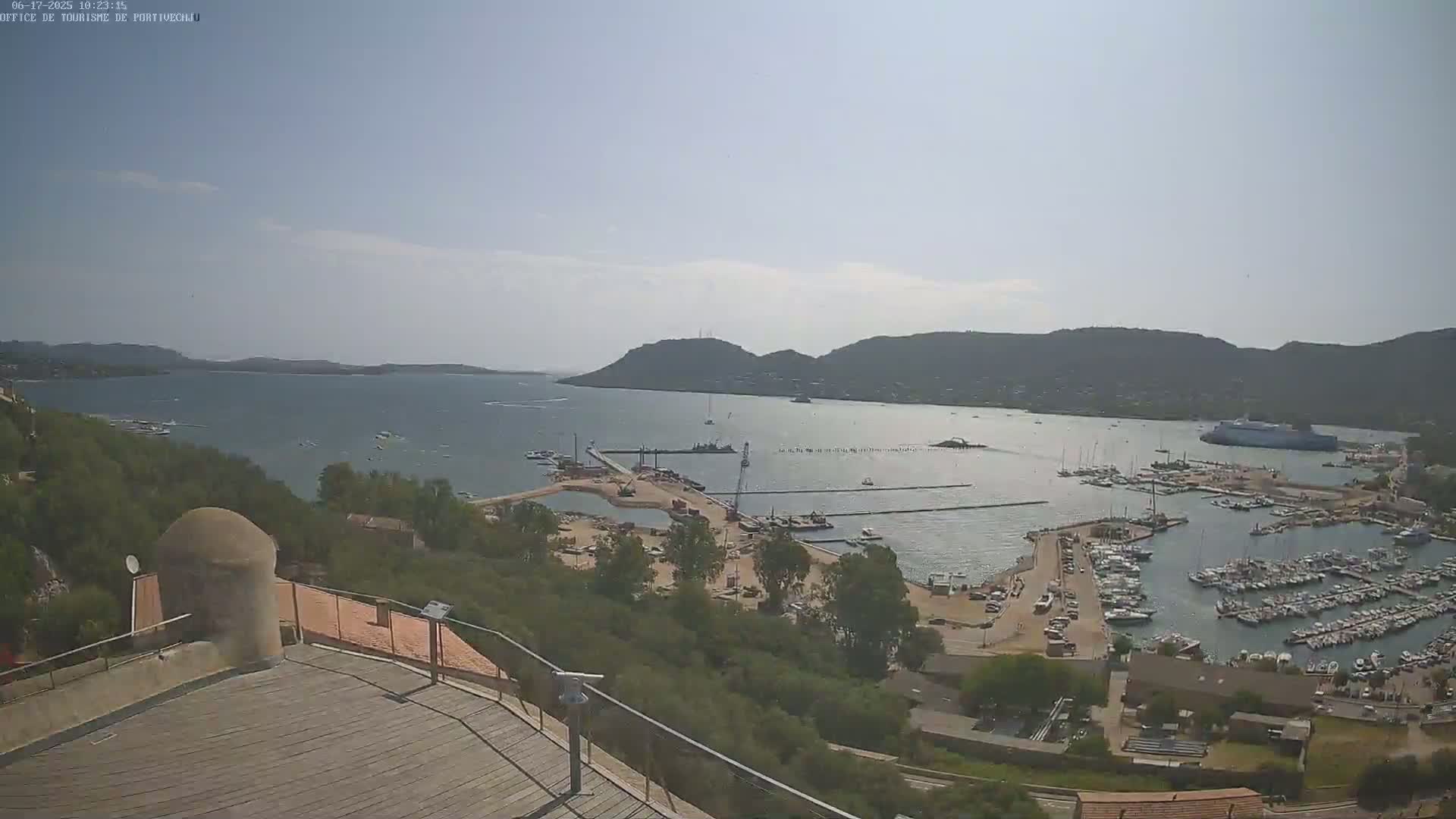 An aerial view of a harbor filled with numerous boats and yachts on a partly sunny day, with a large body of water and hills in the background.
