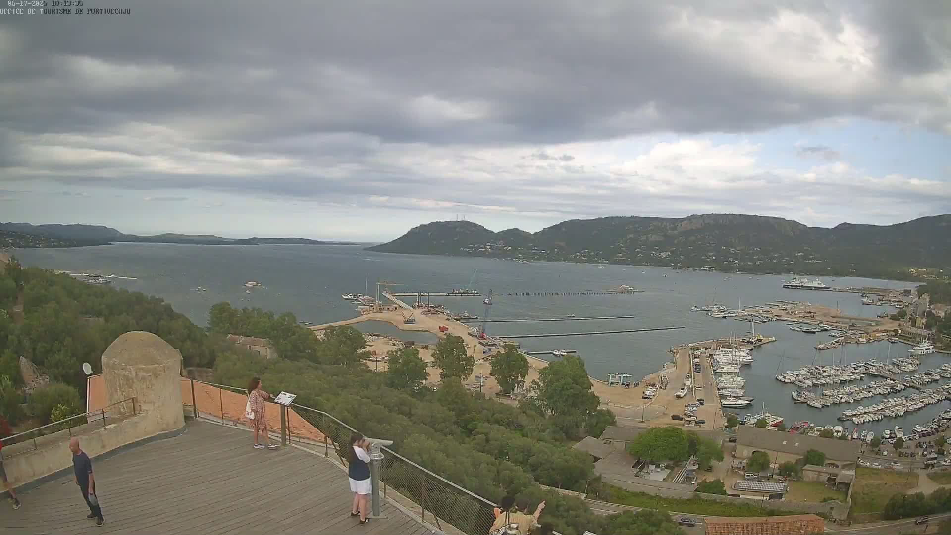 An elevated view of a harbor filled with numerous boats, under a cloudy sky, shows a coastal town nestled amongst hills.