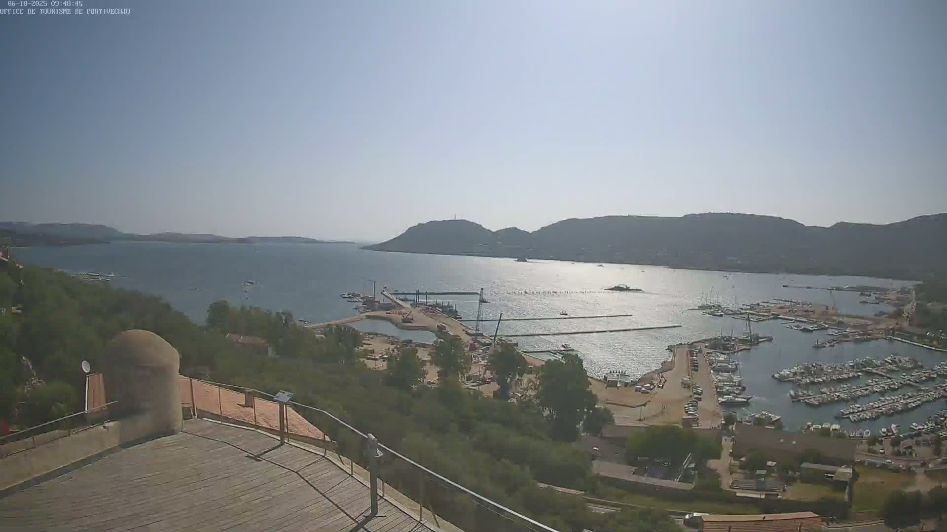 A sunny, high-angle view shows a calm bay with numerous boats docked at a marina, surrounded by green hills and a partially visible town.