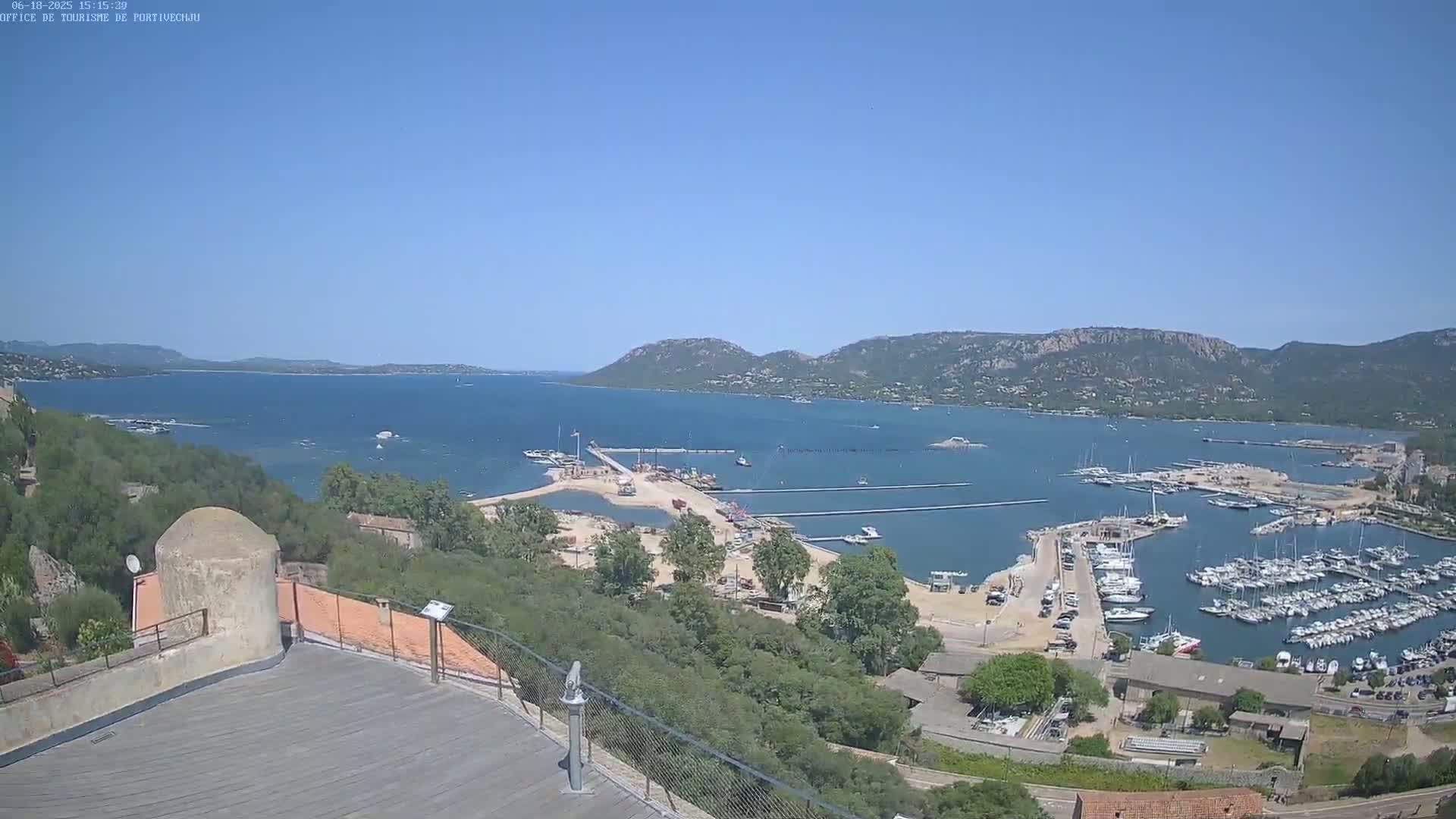 A high-angle, long shot on a clear, sunny day shows a harbor filled with boats and a sandy area under construction, all nestled into a bay surrounded by green hills and a calm blue sea.