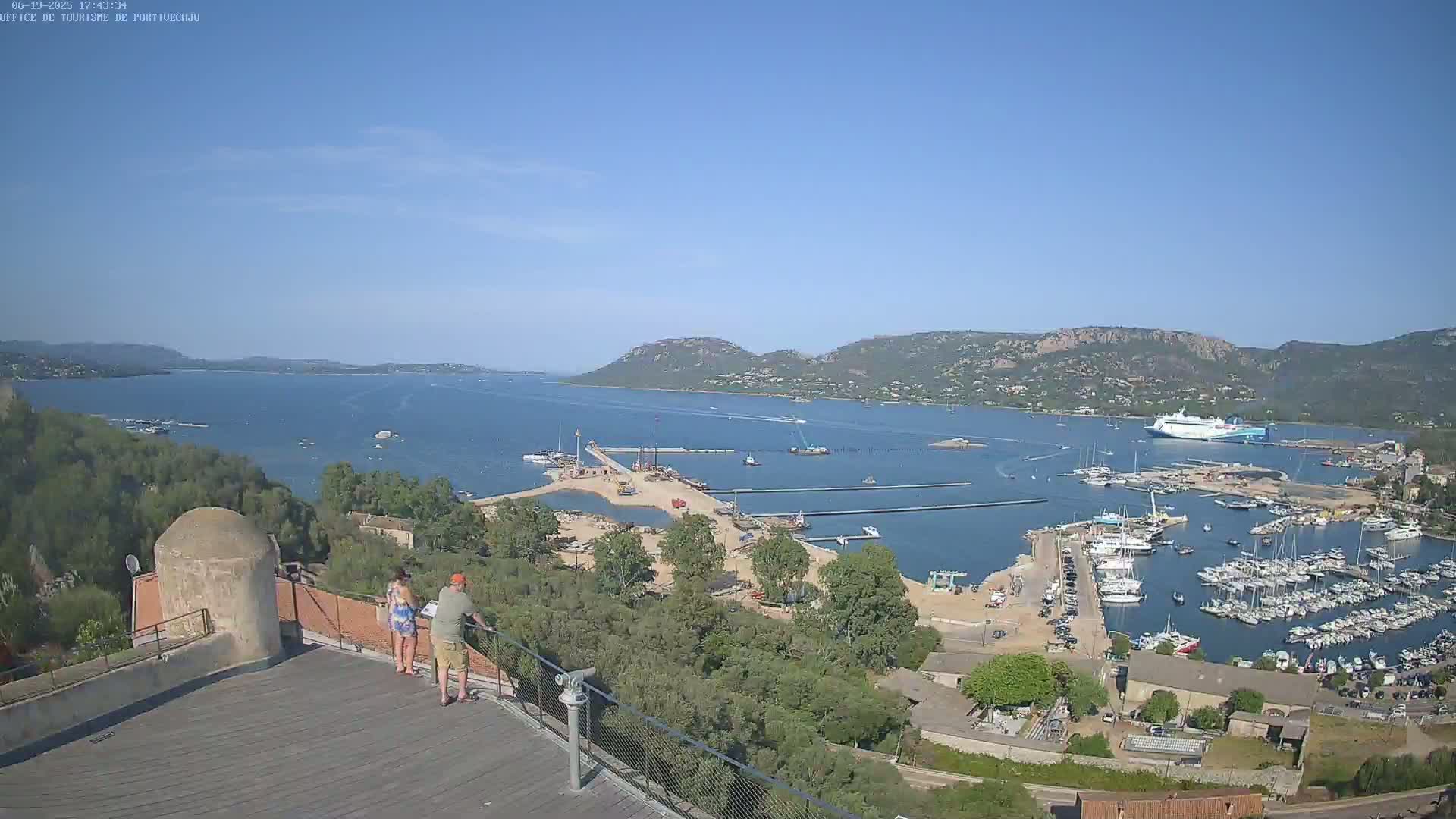 A high-angle view on a clear, sunny day shows a harbor filled with boats, a construction site, and two people standing on a rooftop overlooking the water and distant hills.