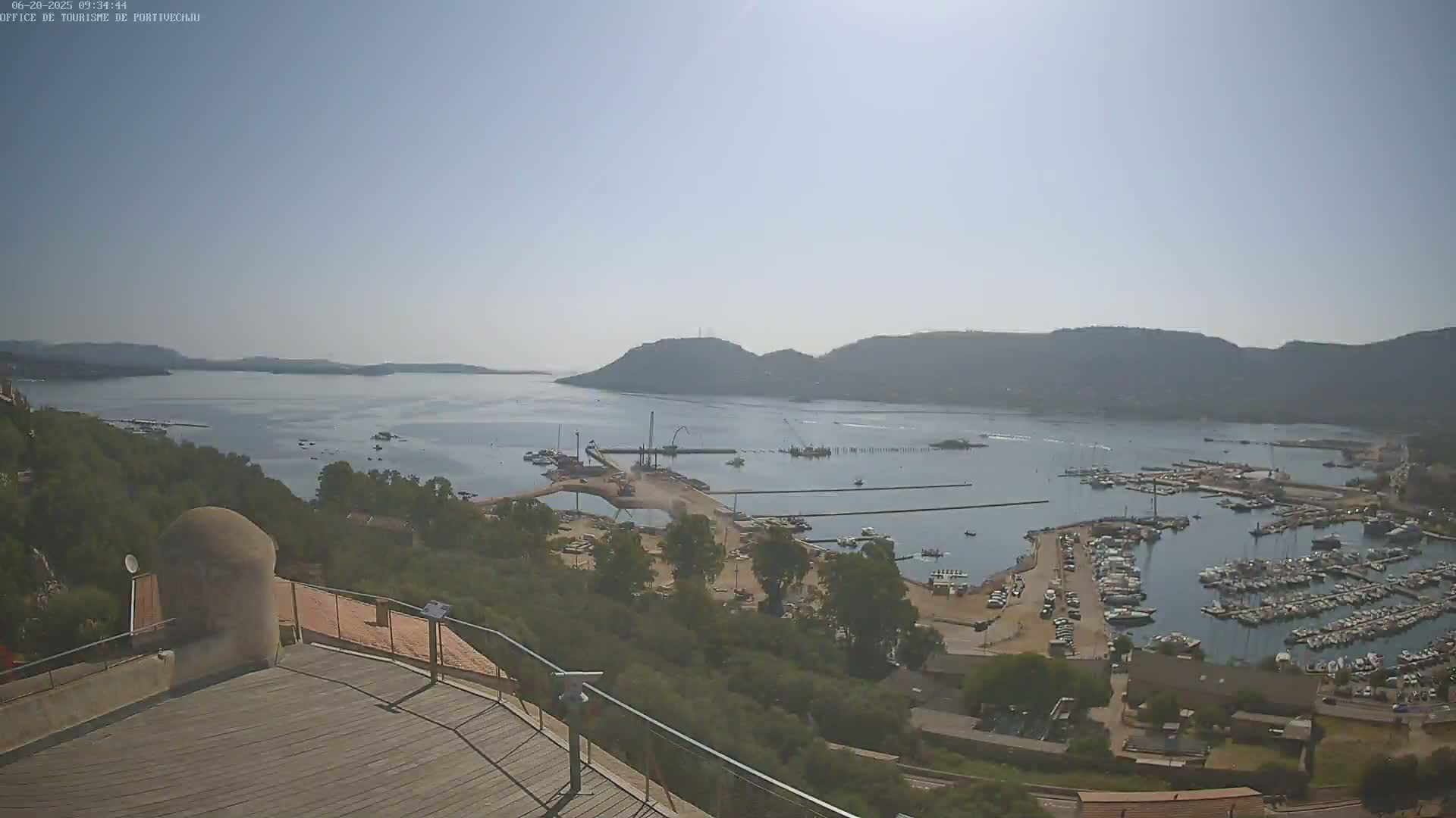 A high-angle, long shot on a sunny day shows a marina filled with boats at the base of a hill overlooking a calm bay and distant mountains.