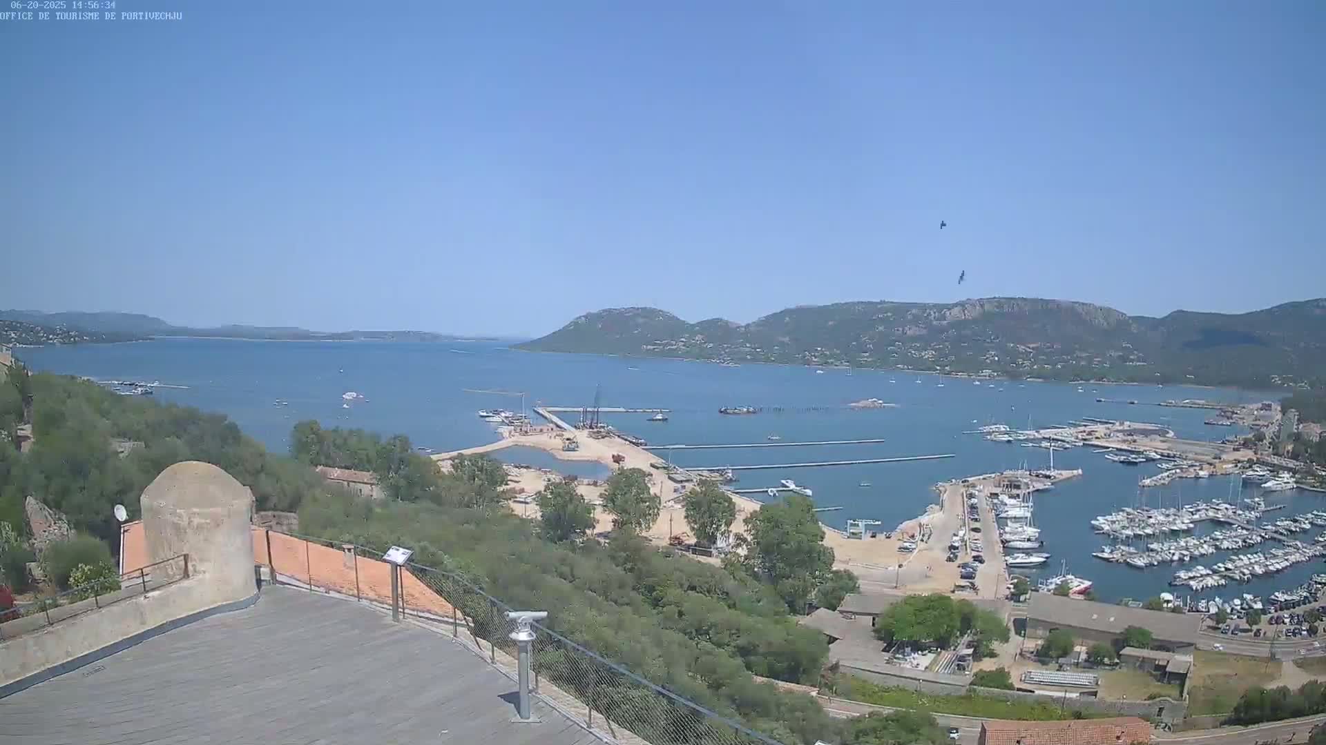 A high-angle, sunny view shows a harbor filled with boats, a sandy area with construction, and a body of water extending to hills under a clear blue sky.