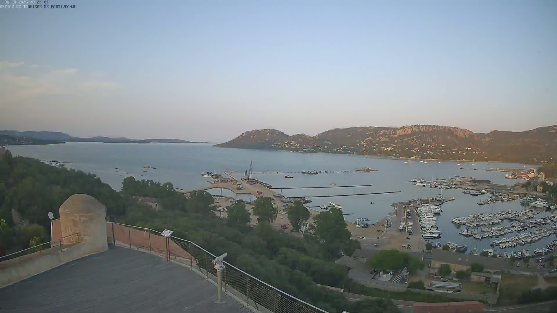 A high-angle, long shot shows a calm bay with numerous boats docked in a marina, surrounded by green hills under a clear, light-blue sky.