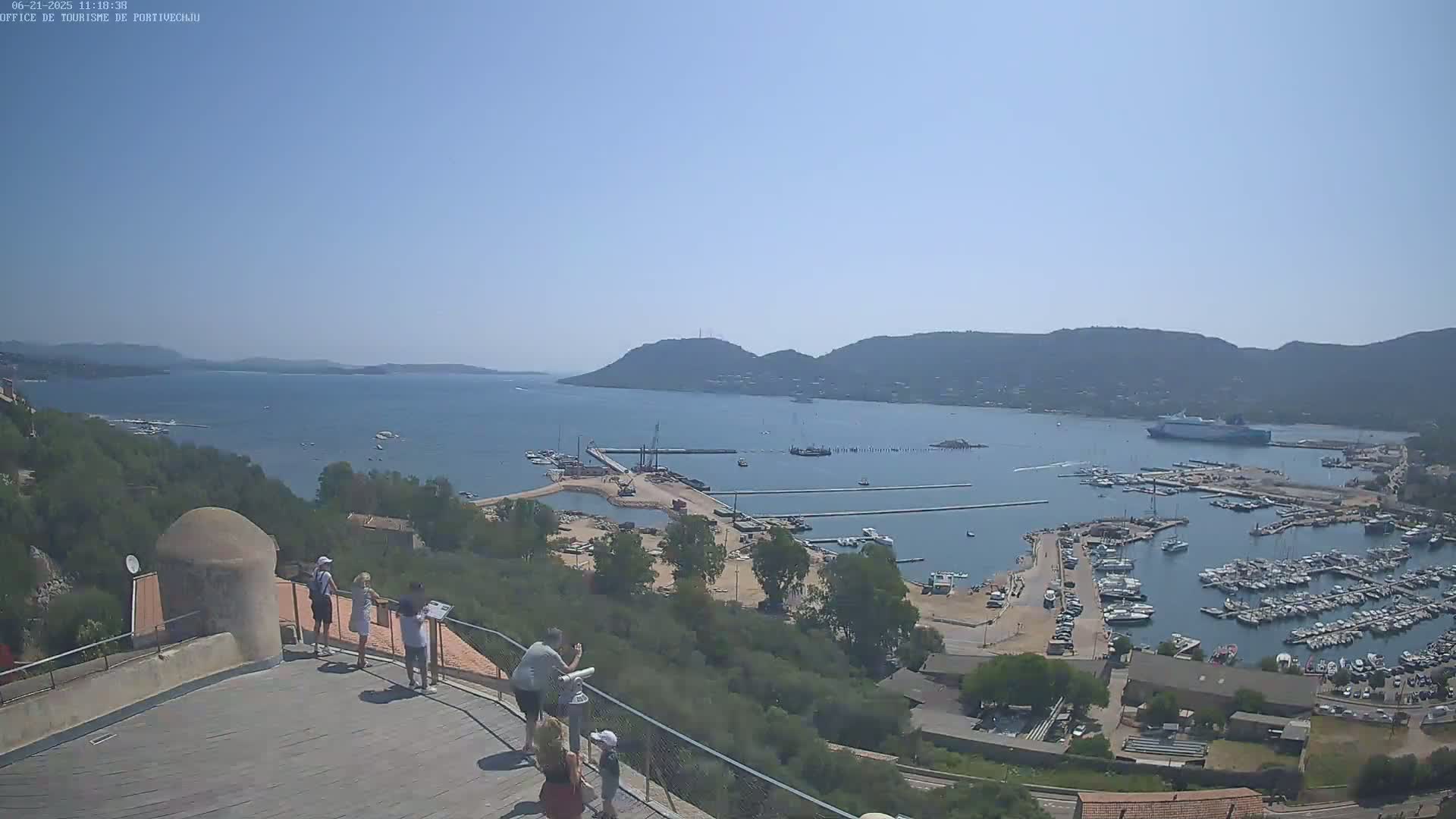 A sunny, clear day overlooks a harbor filled with boats and yachts, situated between hills and a calm body of water, where several people stand on a viewing platform.