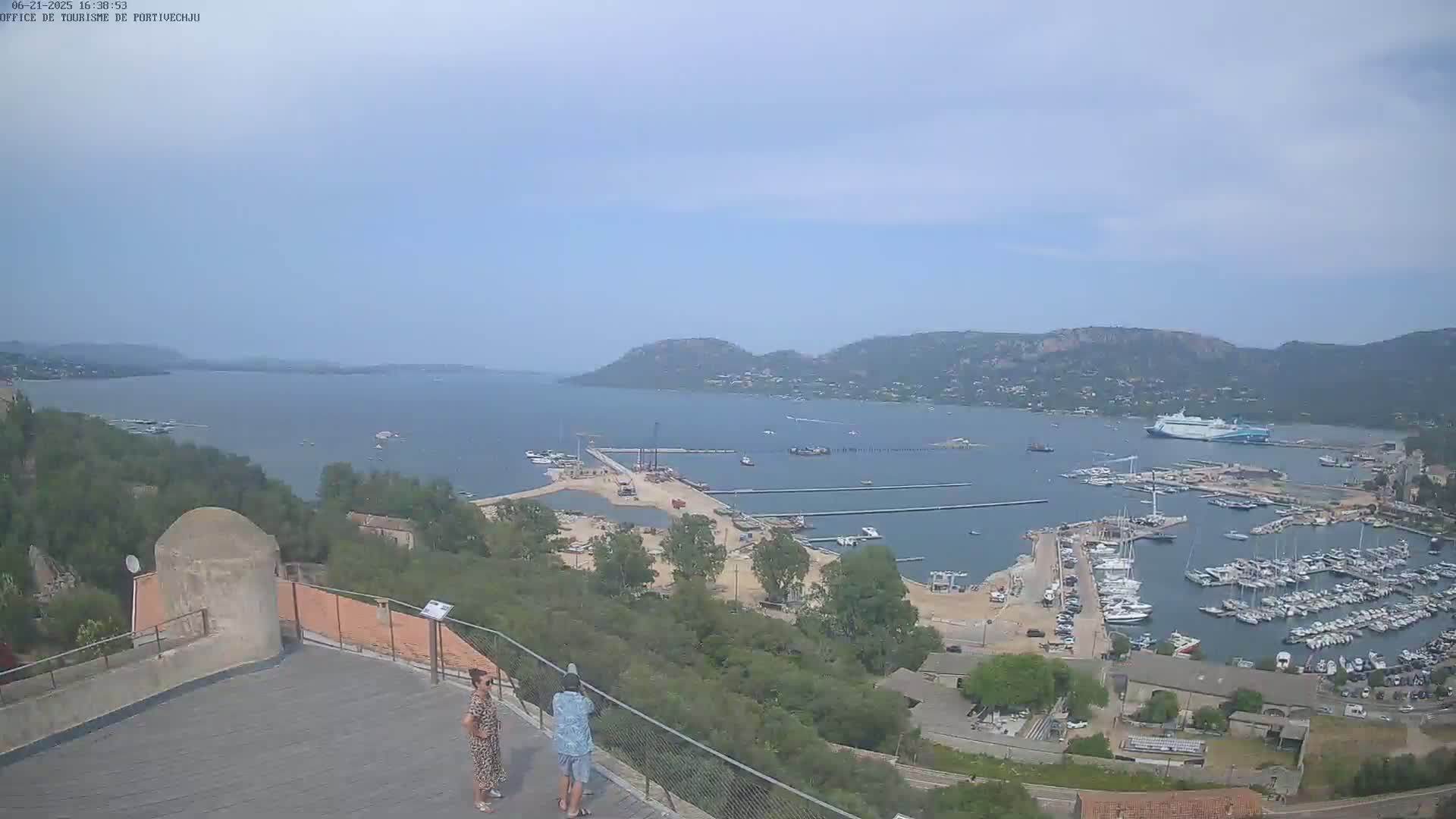 A high-angle, long shot shows a harbor filled with boats and ships on a partly sunny day, with two people standing on a viewing platform overlooking the water and hills in the distance.
