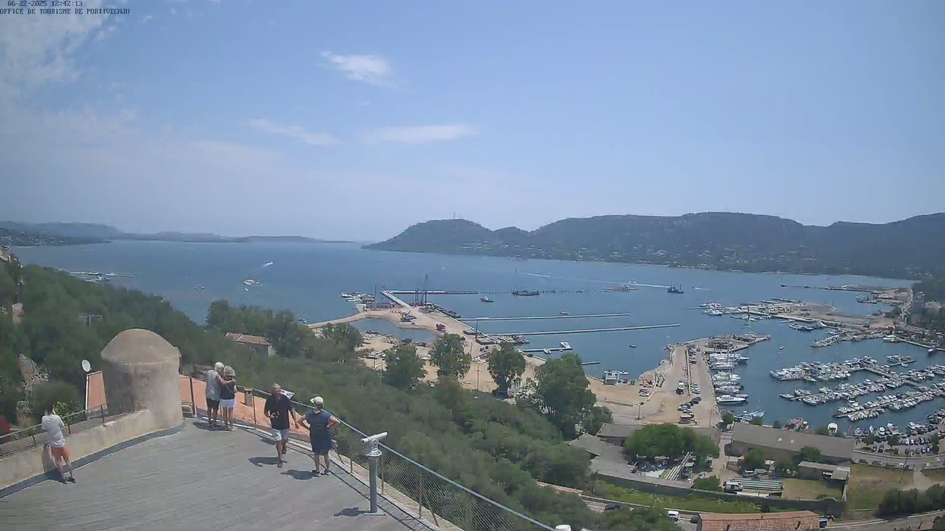 A high-angle, sunny view shows a harbor filled with boats, surrounded by green hills and a calm bay under a mostly clear sky.
