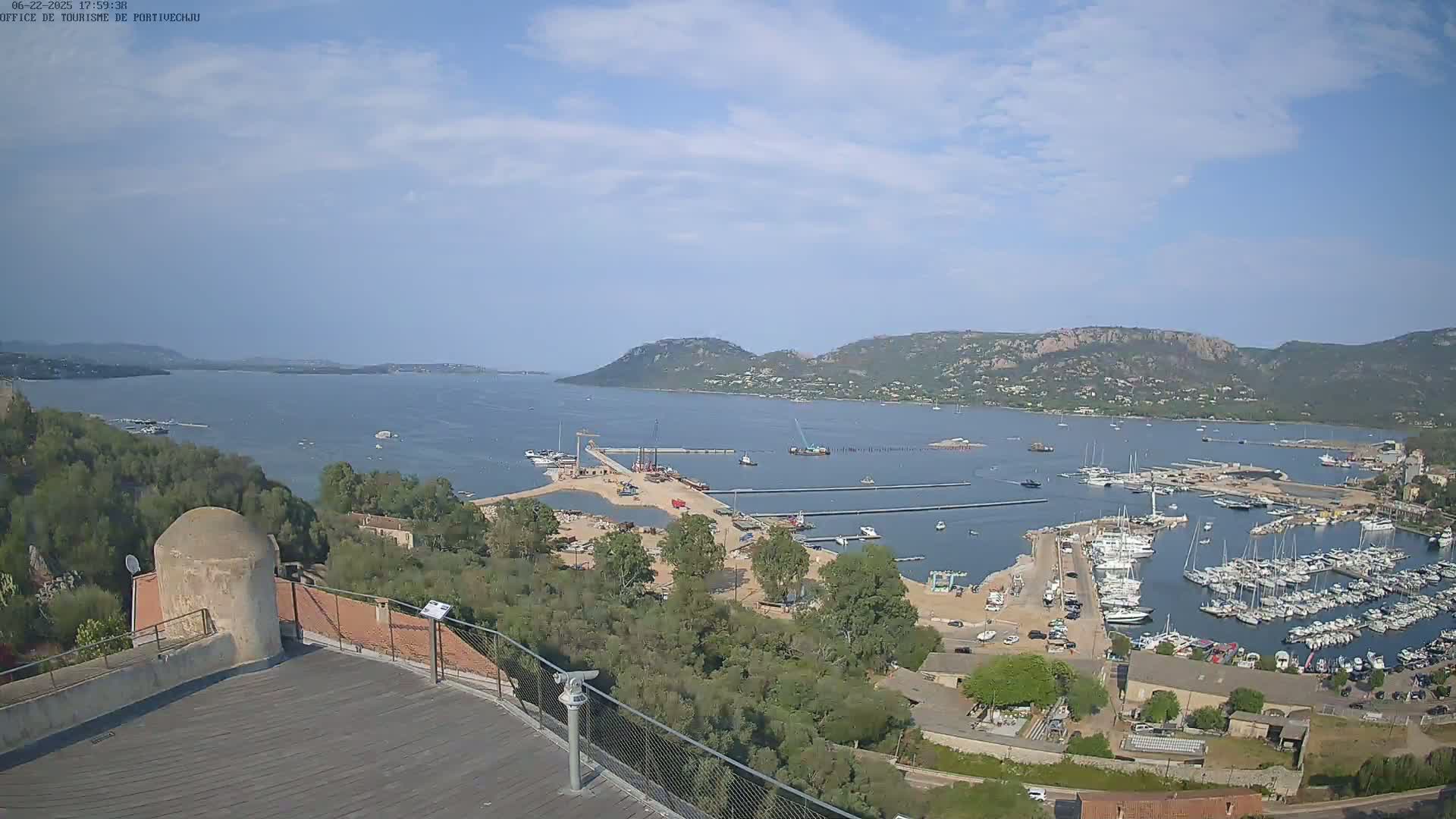 An elevated view shows a harbor filled with boats on a sunny day, with a calm body of water extending to hills in the distance.