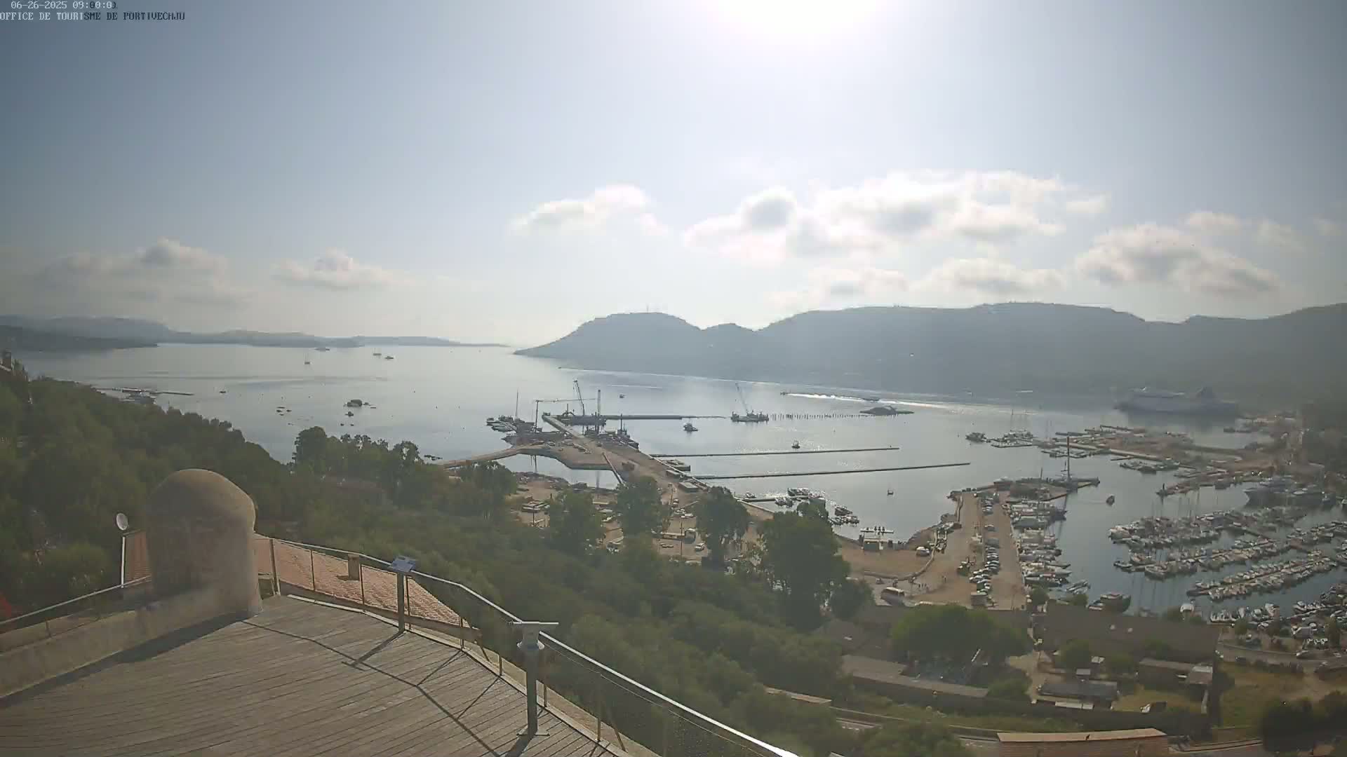 A sunny day reveals a harbor filled with boats, a construction site on a pier, and rolling hills in the background.