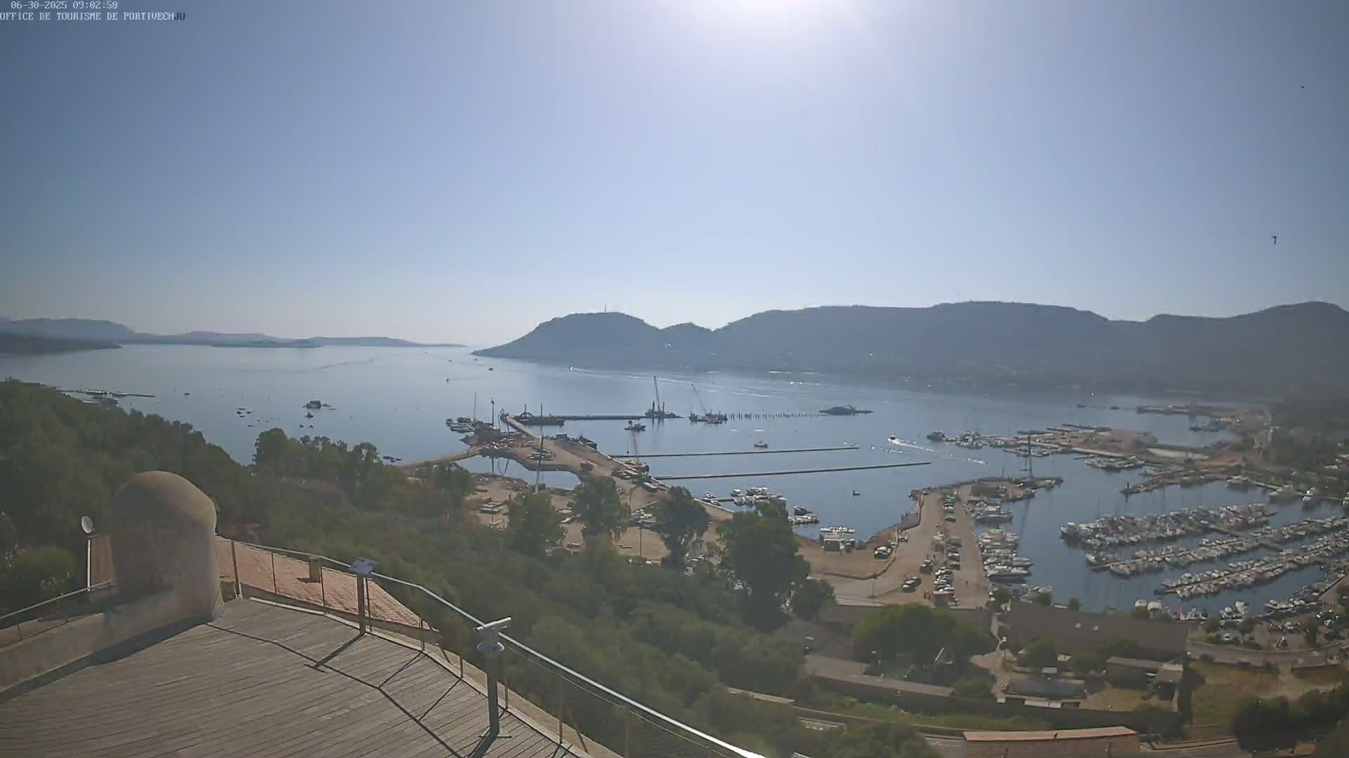 A sunny day overlooking a calm bay with a marina filled with boats, construction activity near the harbor, and hills in the distance.