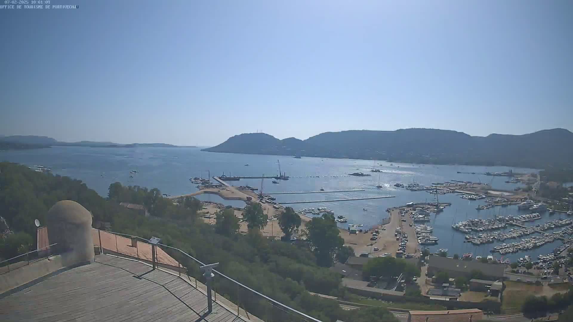 A high-angle view on a clear, sunny day shows a marina filled with boats, surrounded by hills and a calm body of water.