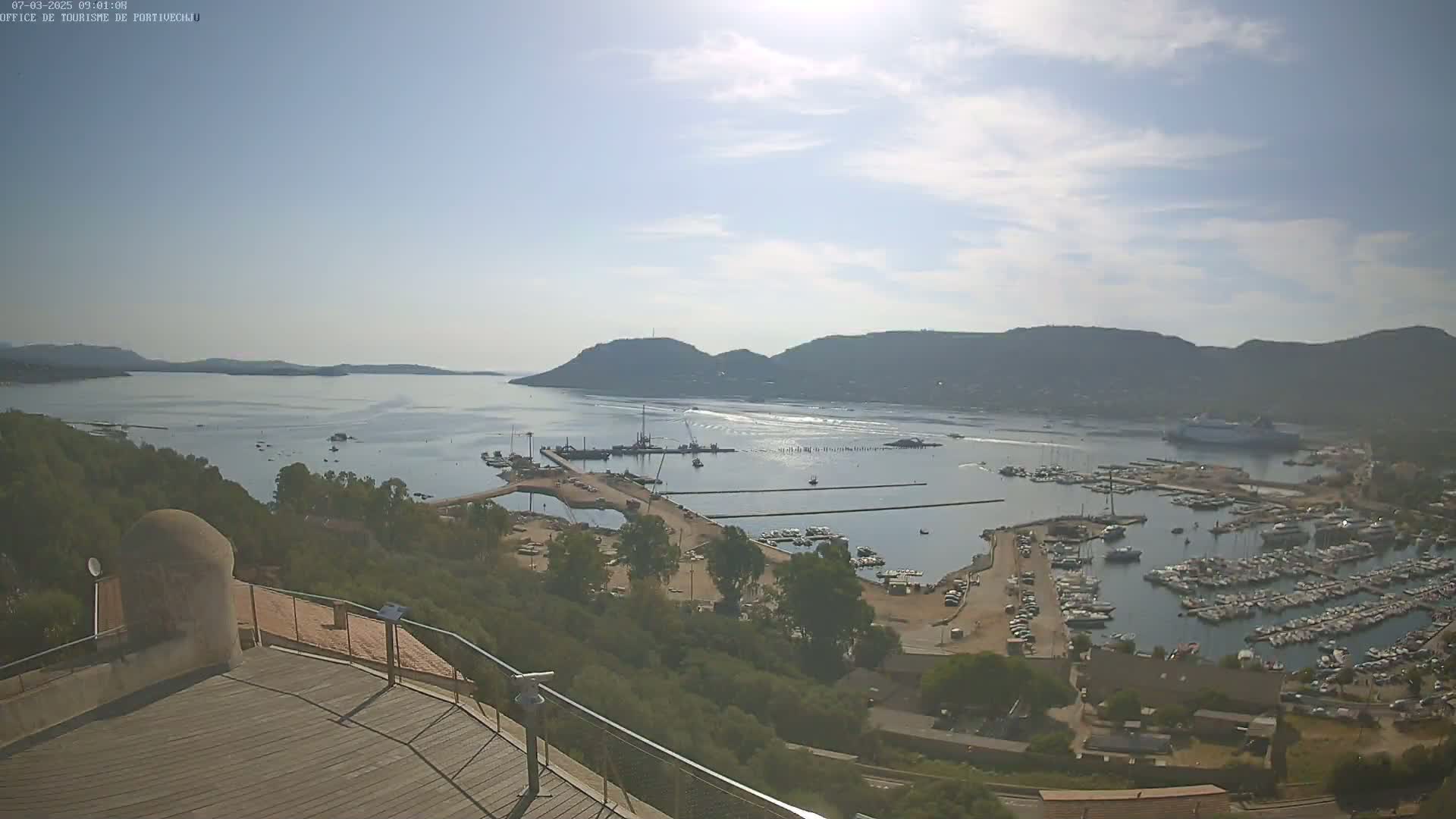 A sunny day reveals a harbor filled with numerous boats and yachts, nestled between a hillside and a calm bay under a partly cloudy sky.