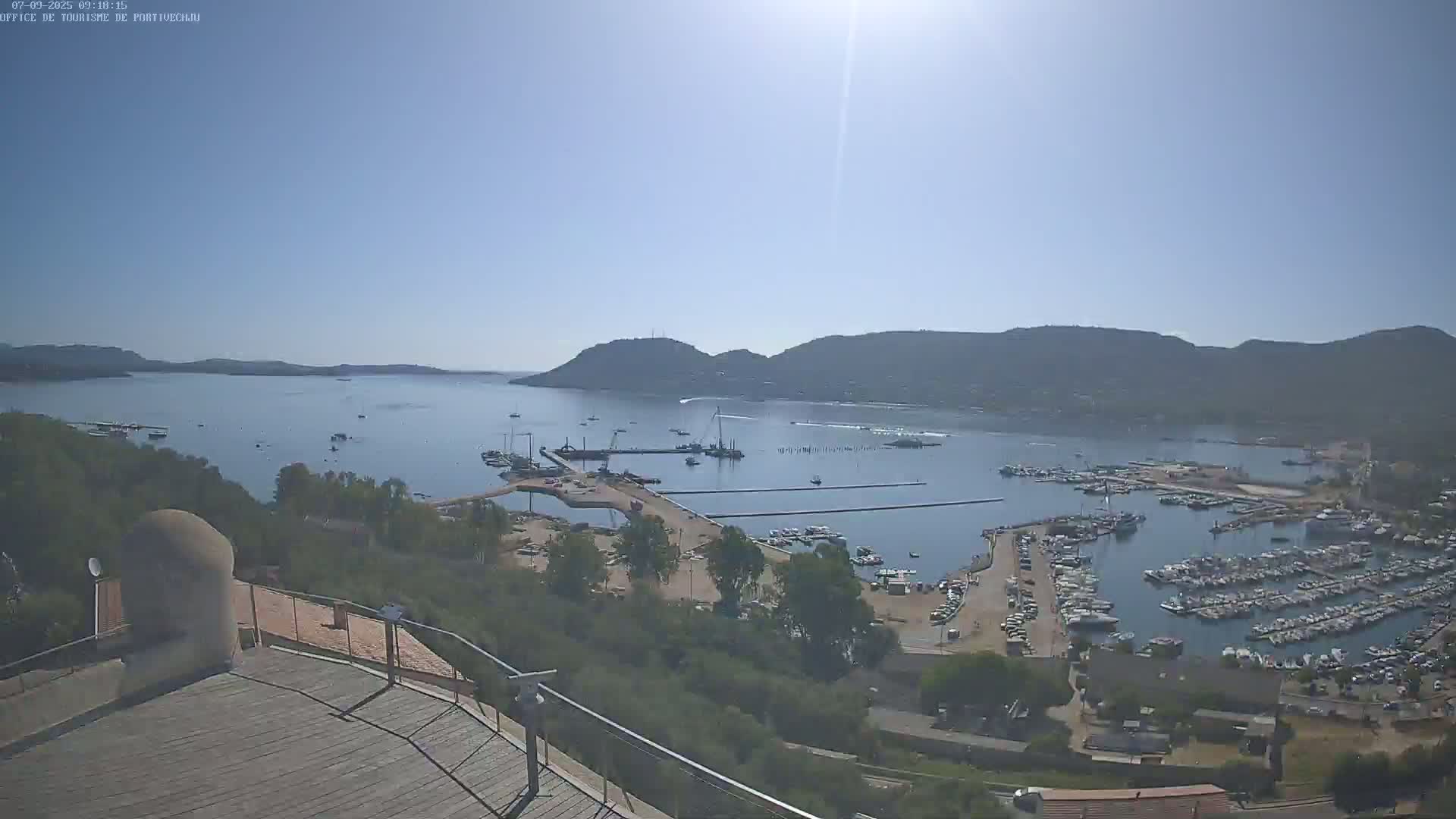 A high-angle, long shot on a sunny day shows a harbor filled with boats, surrounded by hills and greenery, with construction visible in the foreground.