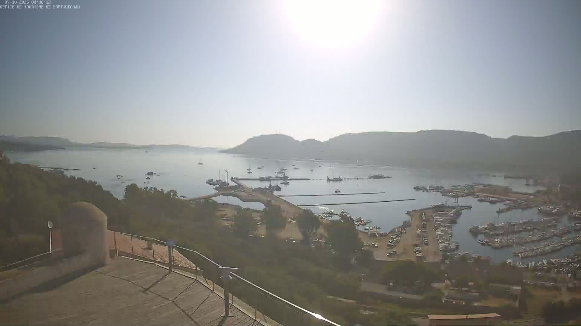 A sunny, hazy day overlooking a calm bay filled with numerous boats and yachts docked in a marina, with hills visible in the distance.