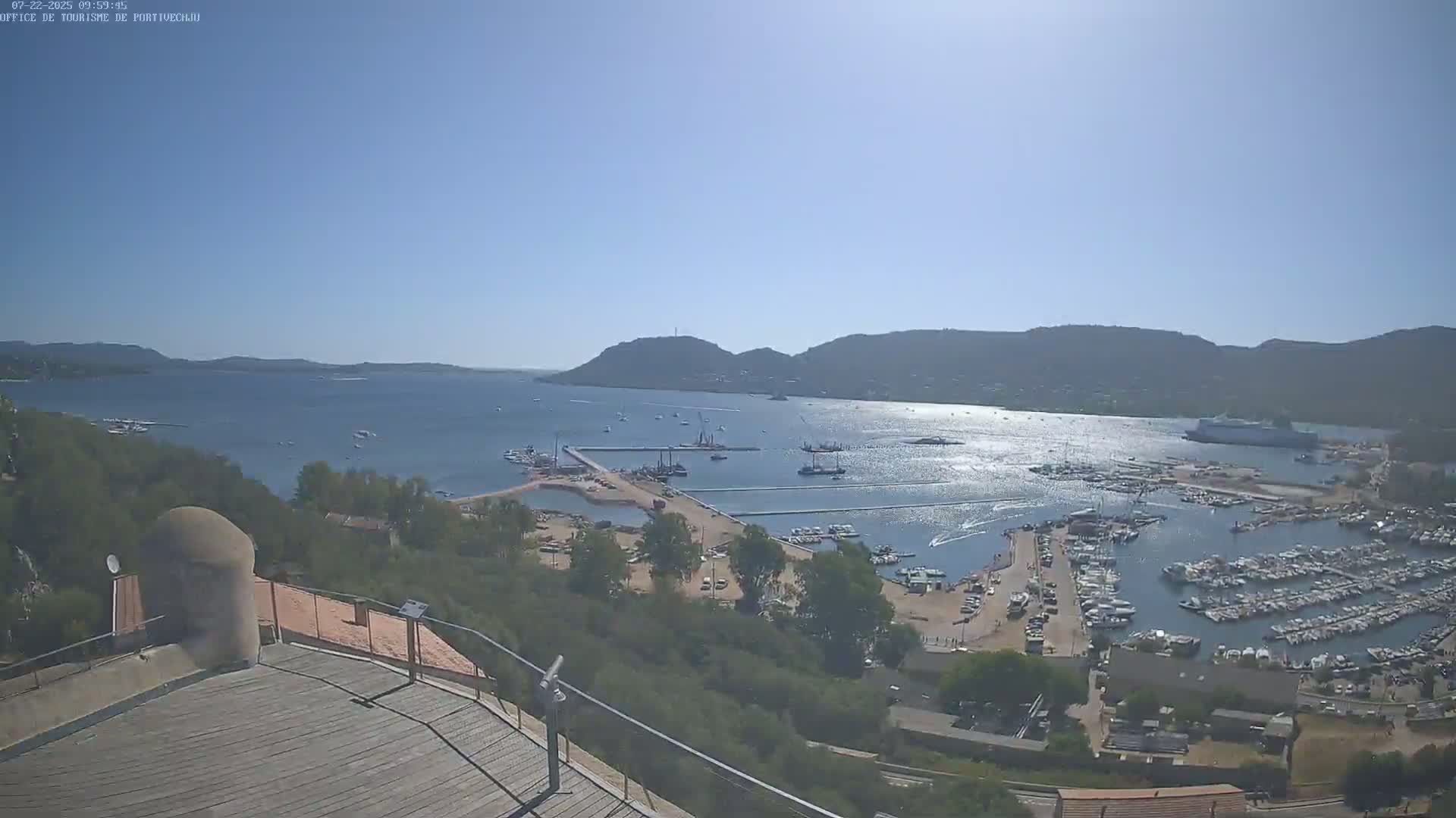 A sunny, high-angle view shows a harbor filled with boats, construction activity, and surrounding hills under a clear blue sky.