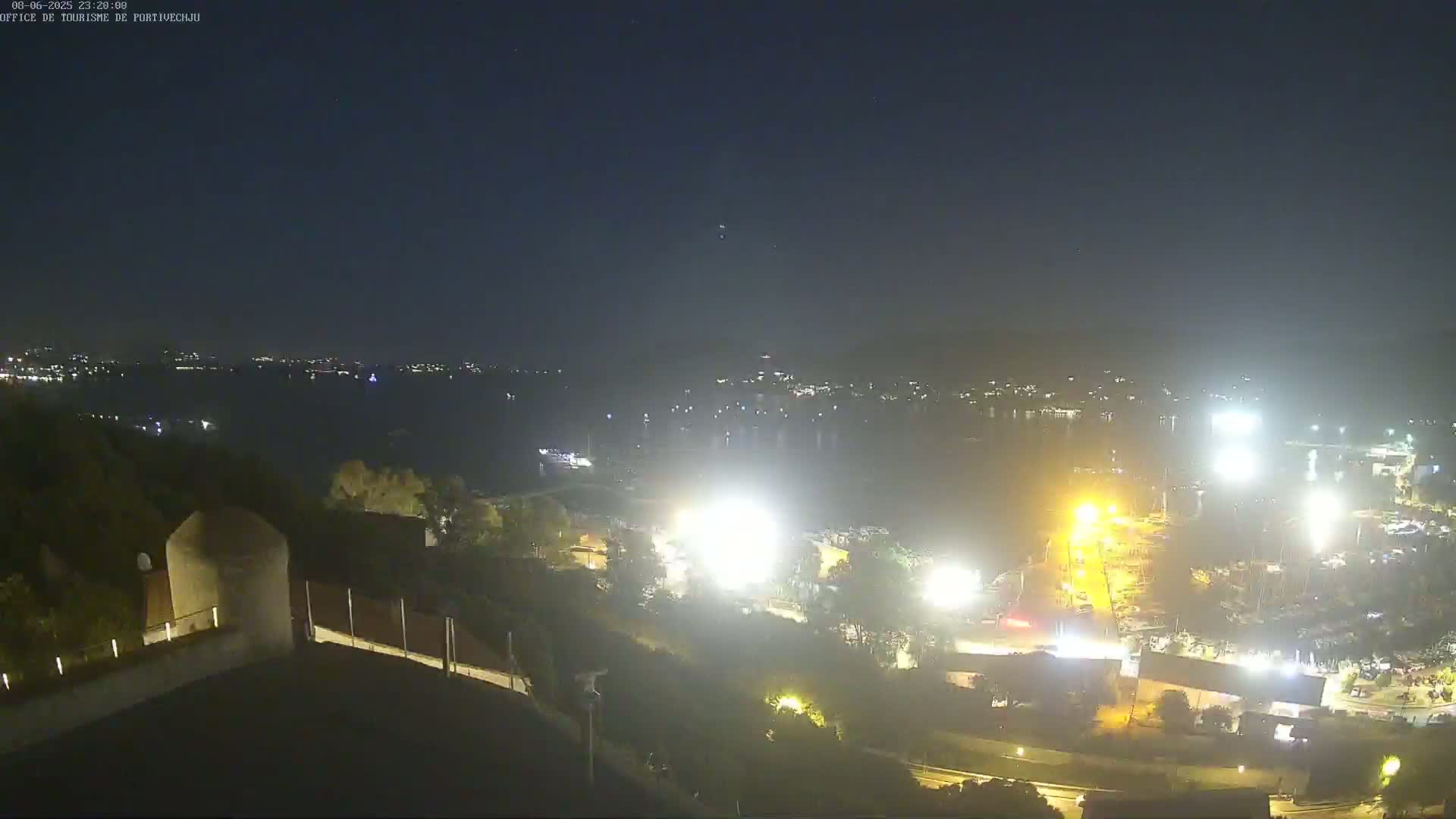 A nighttime, high-angle view shows a body of water with brightly lit boats and buildings along the shore, under a clear, dark sky.