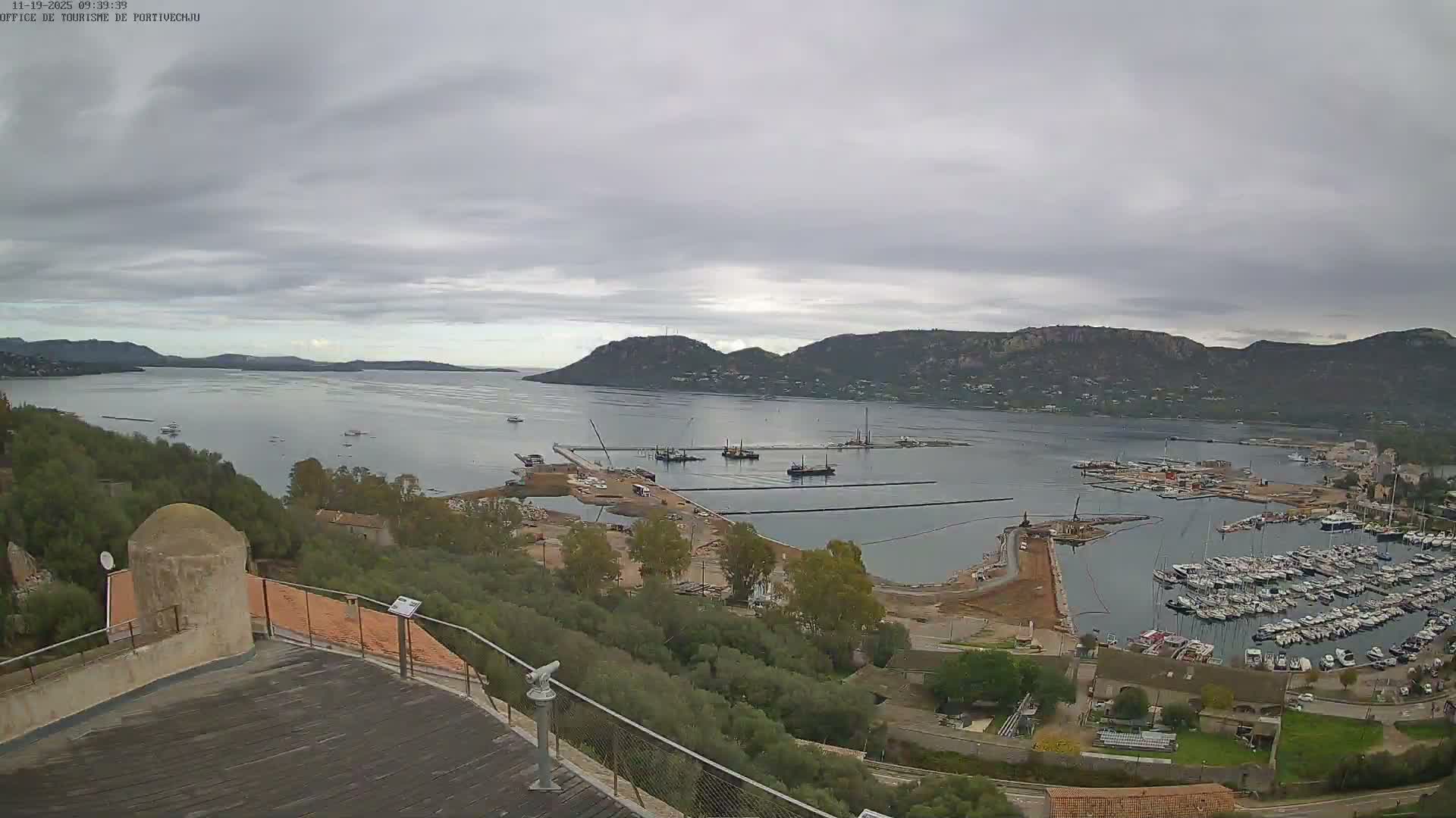 An overcast sky hangs over a panoramic view of a large bay, revealing a bustling marina filled with countless docked boats, active construction along new piers, and distant mountainous shores, with a viewing platform in the foreground.