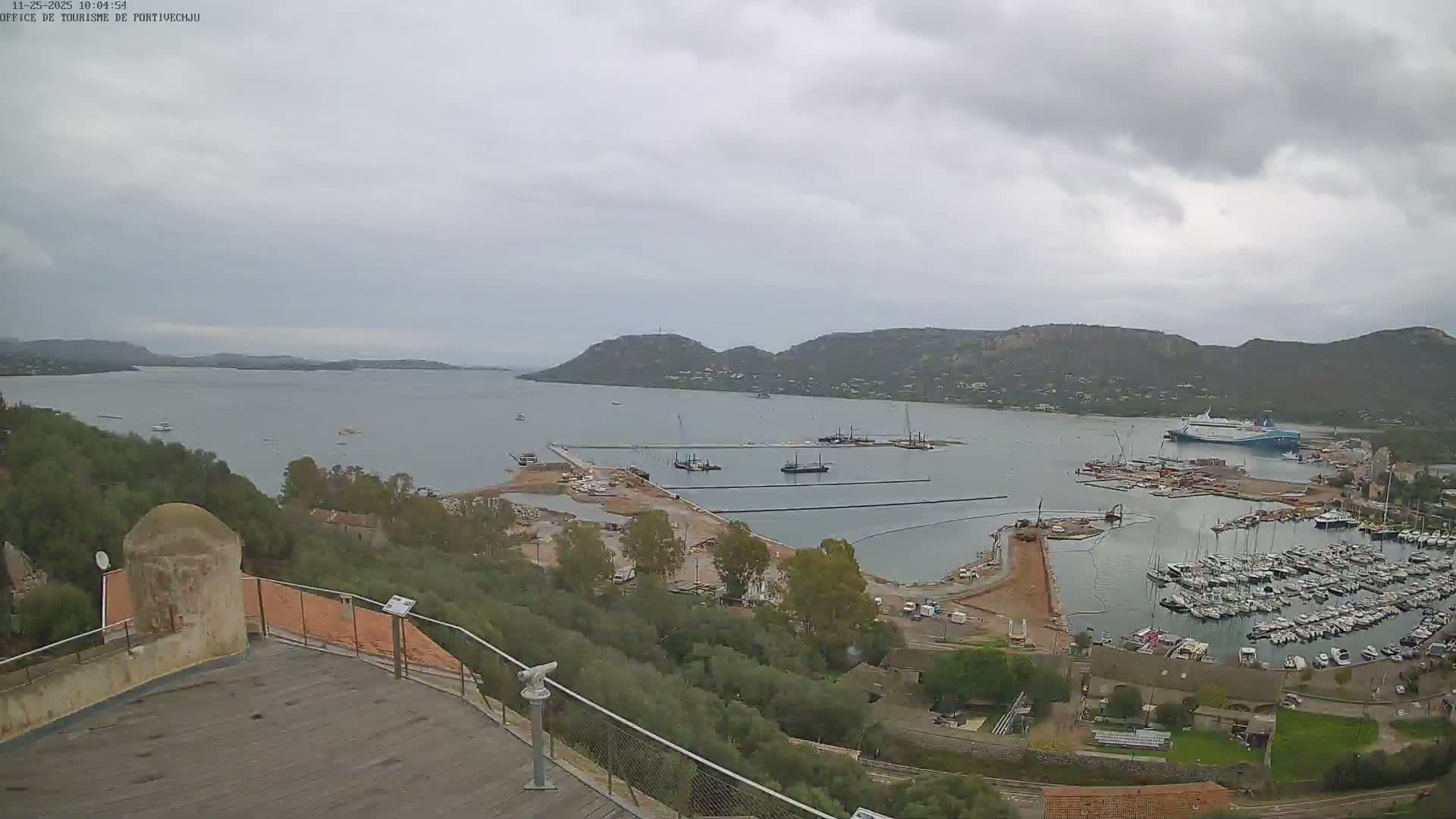 An overcast sky hangs over a bustling harbor filled with numerous docked boats, a large ferry, and ongoing construction, all nestled among hills and viewed from a high observation deck.