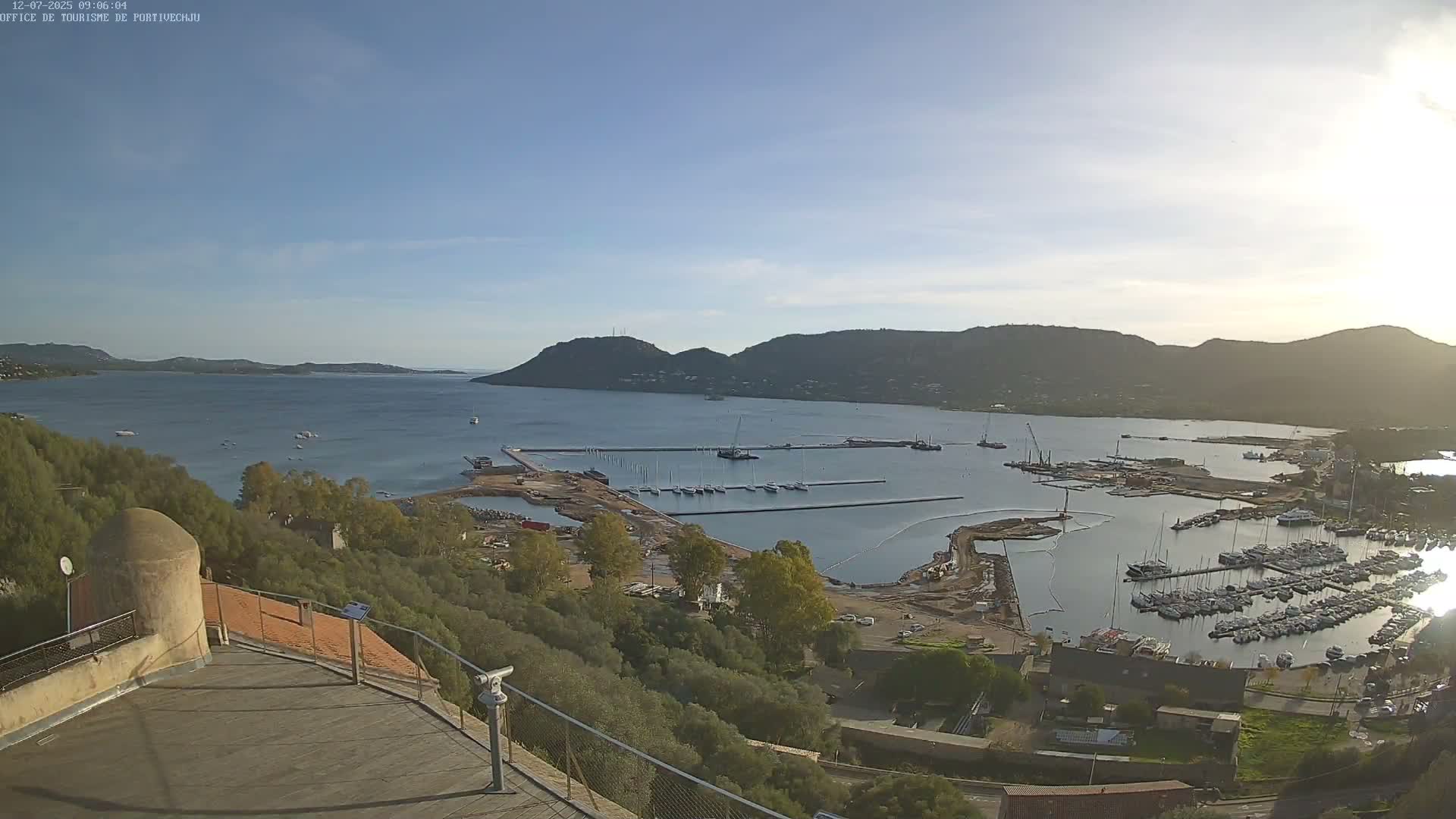 A panoramic view from a high vantage point reveals a bustling port and bay with numerous boats and ongoing construction, surrounded by green hills under a bright, clear, and sunny sky.