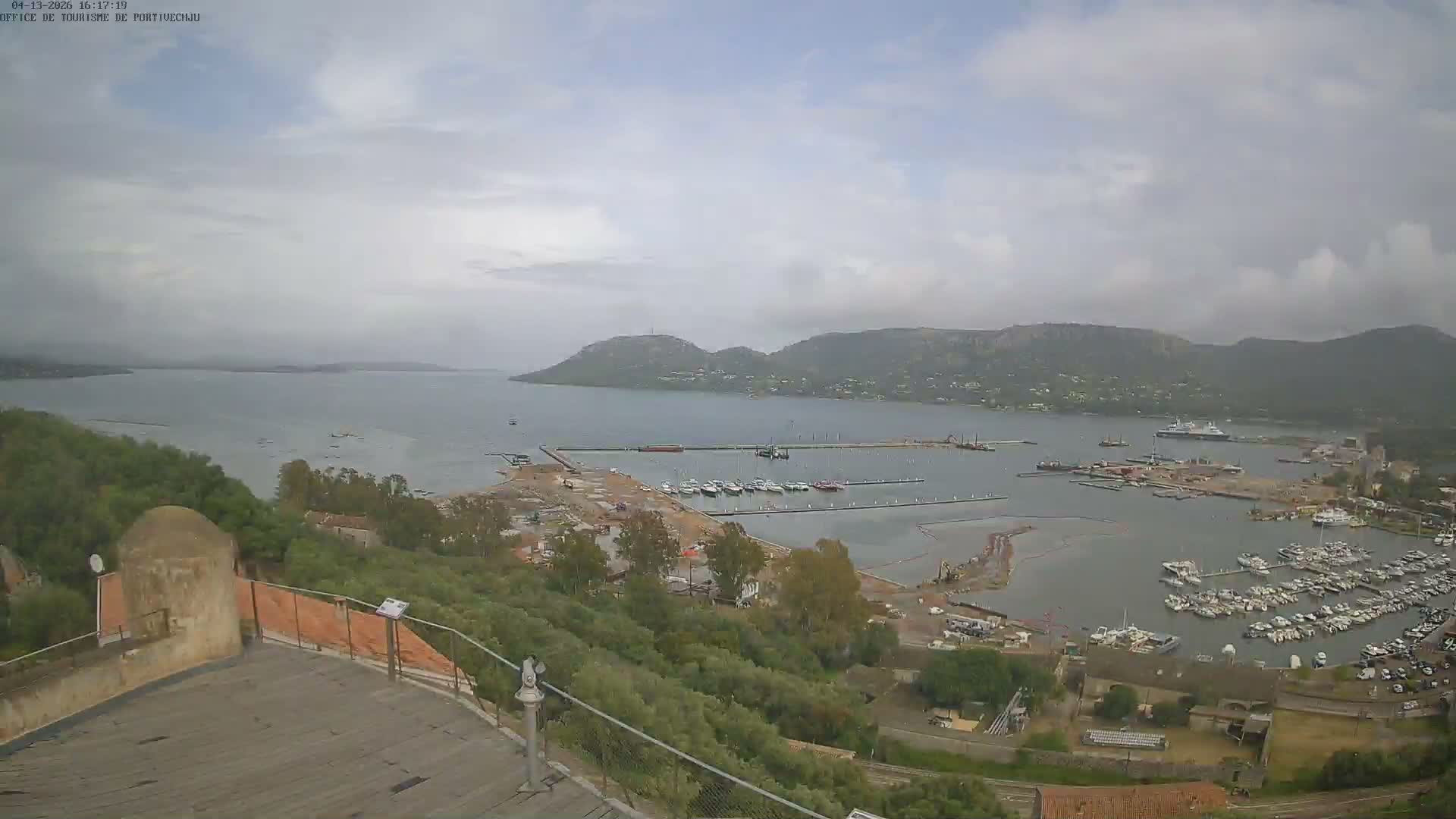 A panoramic view from a high vantage point reveals a bustling port and bay with numerous boats and ongoing construction, surrounded by green hills under a bright, clear, and sunny sky.