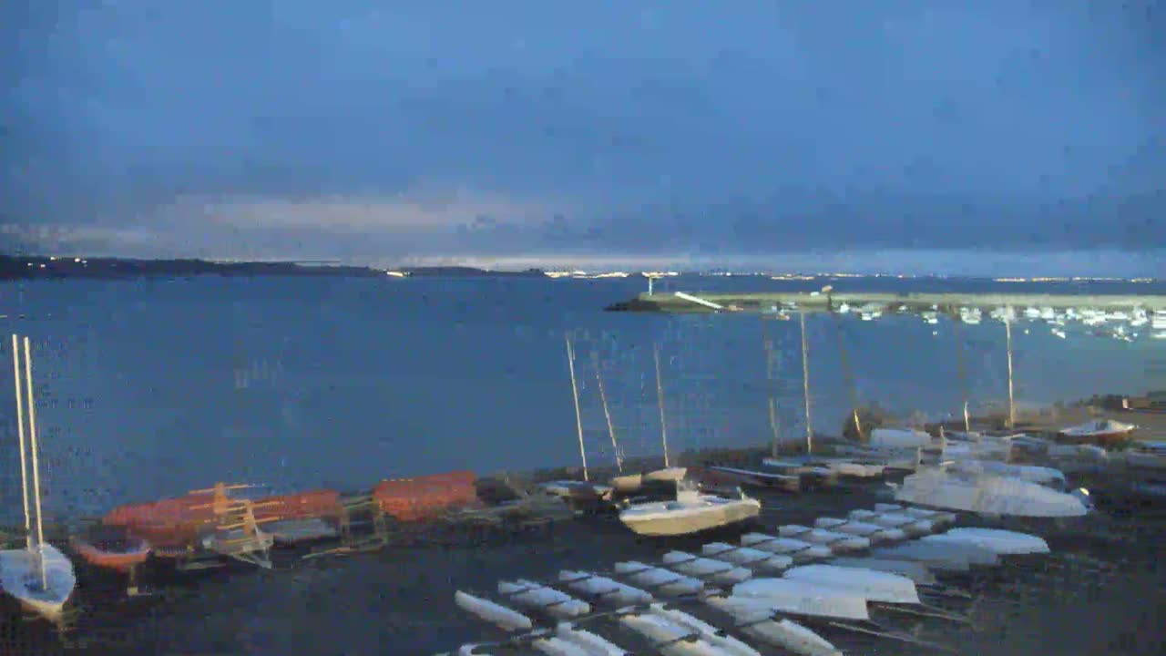 Numerous boats and sailboats are docked along a dark shoreline at twilight, with a calm body of water extending to a distant, faintly lit coastline under a mostly clear, dark blue sky.