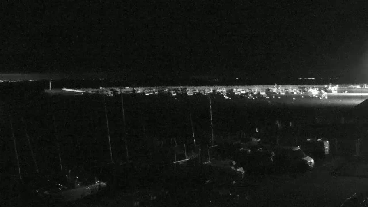 A nighttime, low-light view shows a harbor filled with boats and cars parked in a lot, under a dark sky.