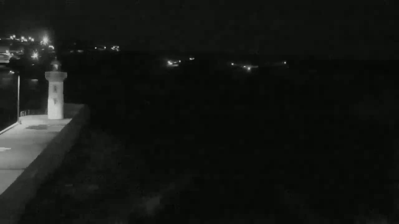 A small, light-colored lighthouse-like structure stands on a dark, nighttime waterfront, with distant, faint lights visible in the background.