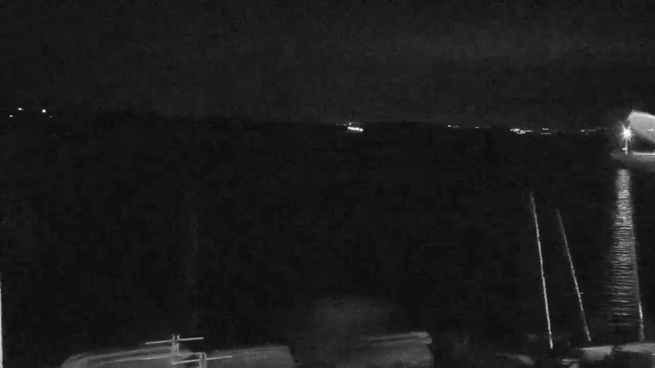 A nighttime, low-light view of a dark body of water with faintly visible lights in the distance and a few vertical lines, possibly dock pilings, near the bottom of the frame.