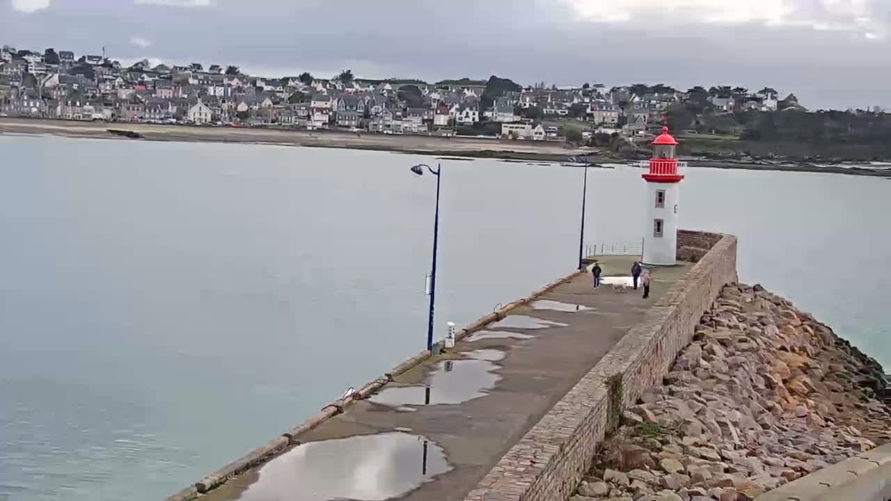 A seaside town is visible across a calm body of water from a wet, stone pier with a lighthouse at the end under an overcast sky.