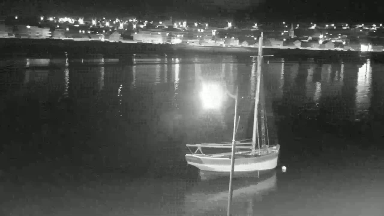 A small sailboat is moored in calm water at night, with a town's lights reflecting on the water in the background.