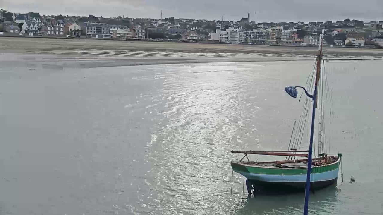 A small sailboat is moored in calm, gray water near a sandy beach and a town under an overcast sky.