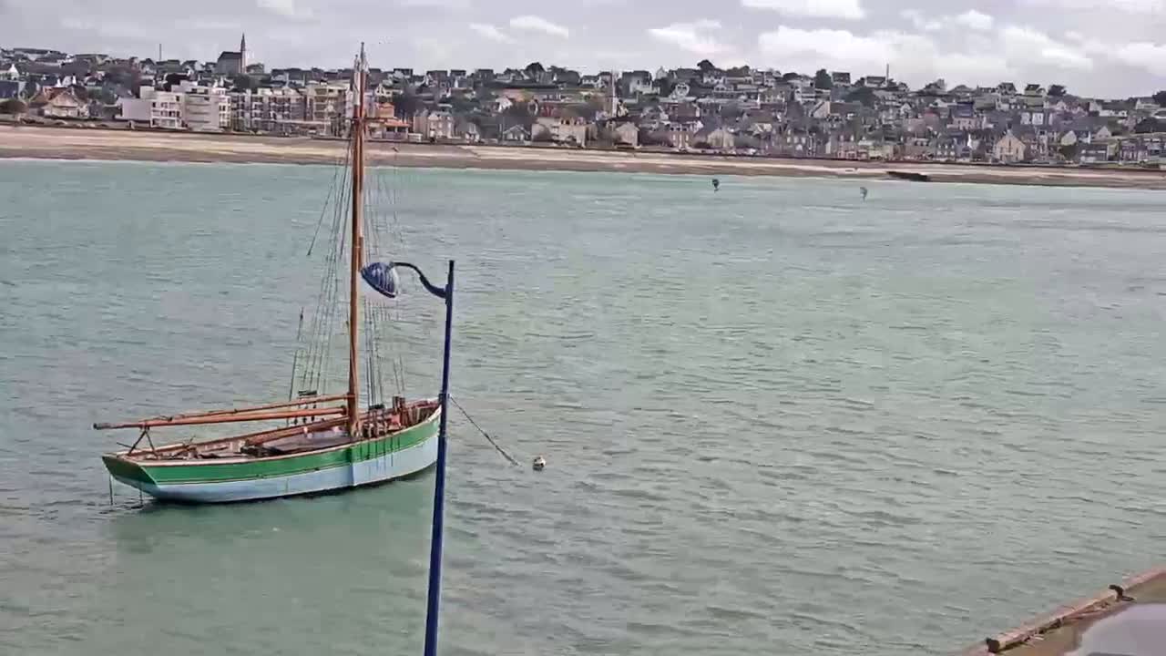 A small sailboat is moored in calm, light-green water in front of a coastal town on an overcast day.