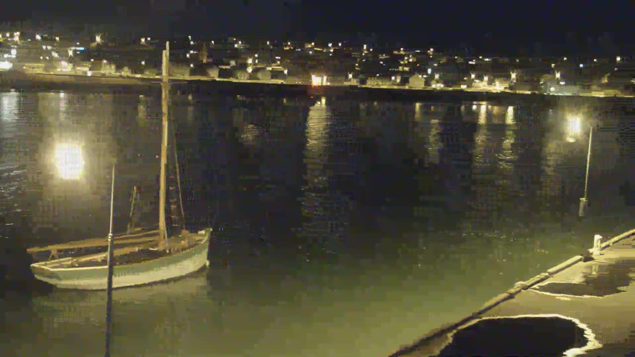 A sailboat is moored at a dock at night next to a calm body of water, with a town lit up in the background.