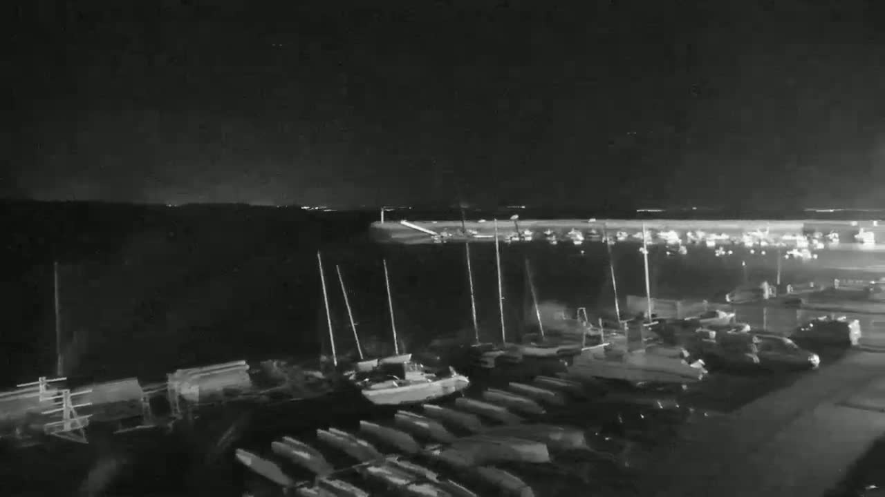 A nighttime, low-light image shows numerous boats docked at a marina, with additional boats and vehicles parked nearby.