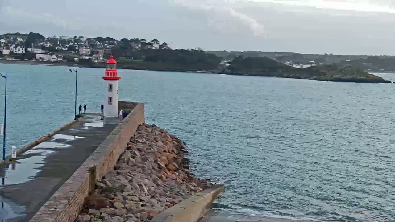 A red and white lighthouse stands on a stone pier overlooking a calm body of water, with a town visible in the distance under a cloudy sky.