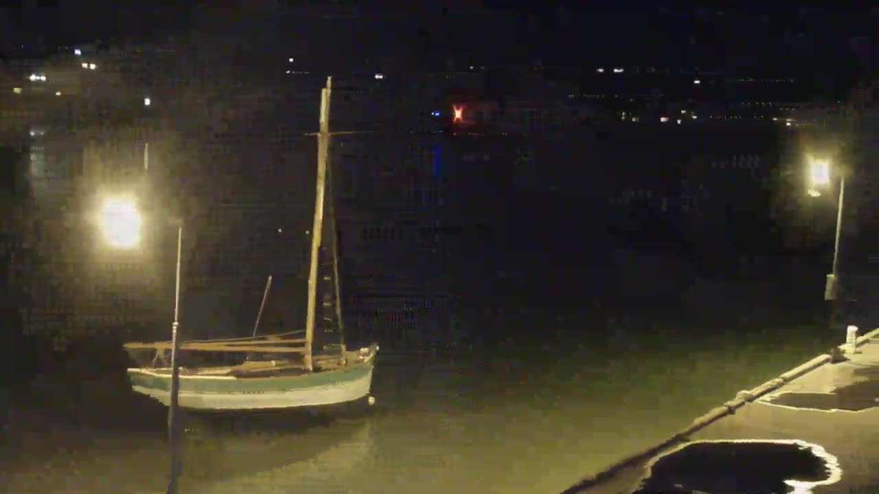 A small sailboat is moored in a dark harbor at night, illuminated by streetlights.