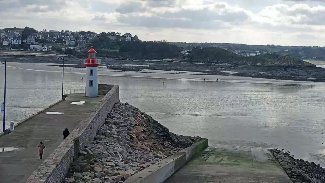 A red and white lighthouse stands on a stone jetty overlooking a calm, gray body of water with a few people visible on the exposed seabed in the distance under a cloudy sky.