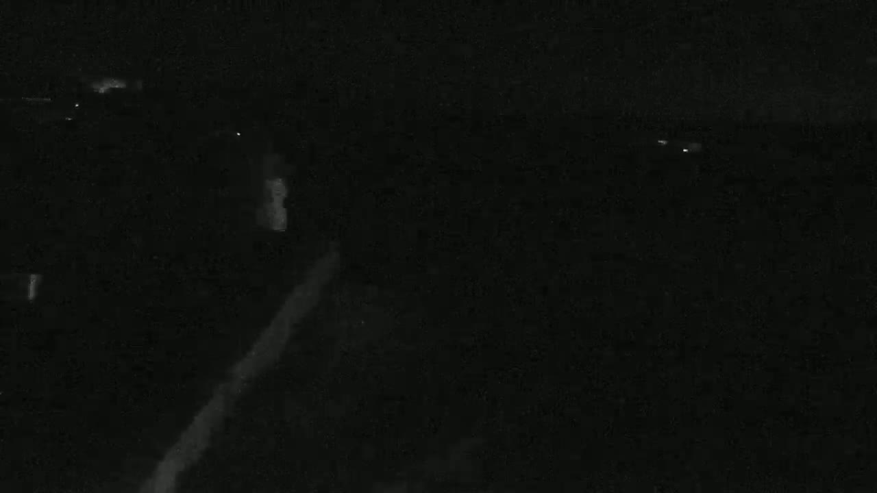 A low-light nighttime image shows a dark landscape with faint, indistinct shapes and a few small points of light in the distance.
