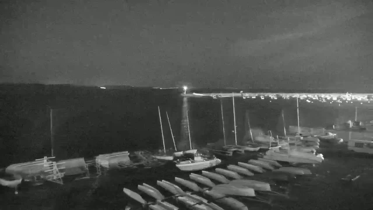 A nighttime, low-light view of a harbor filled with docked sailboats and other small watercraft, under a dark, mostly clear sky.